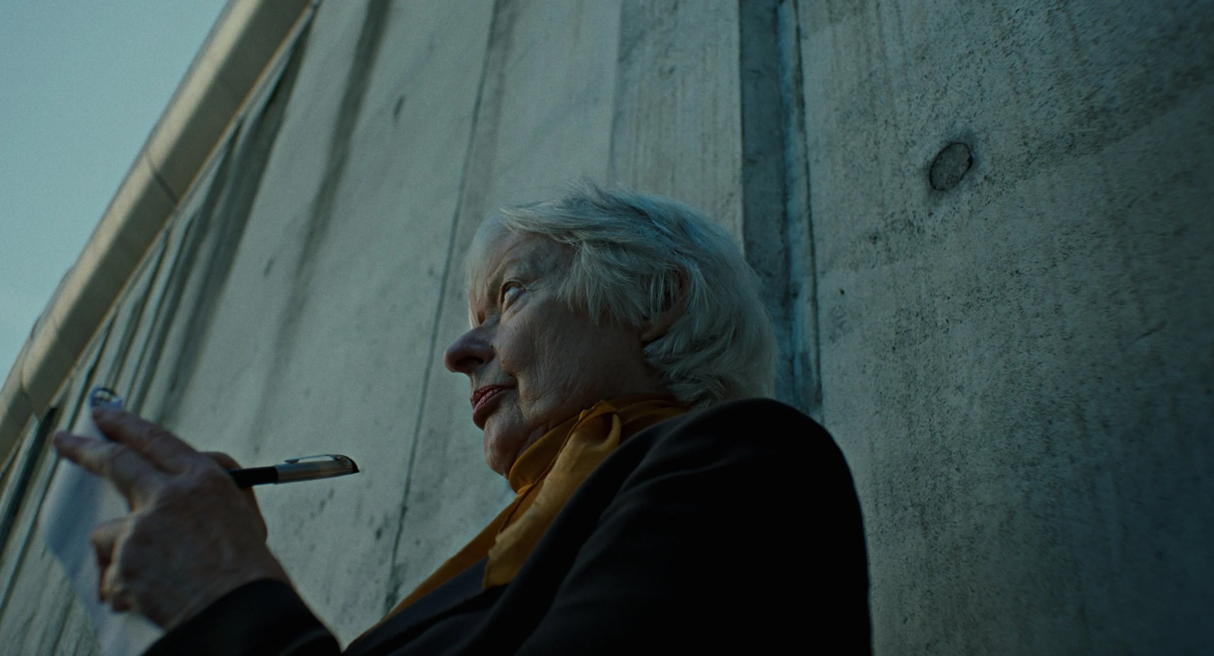 An elderly woman with silver hair, wearing a dark jacket and a yellow scarf, is sitting against a concrete wall, holding a pen in her hand and looking off to the side.