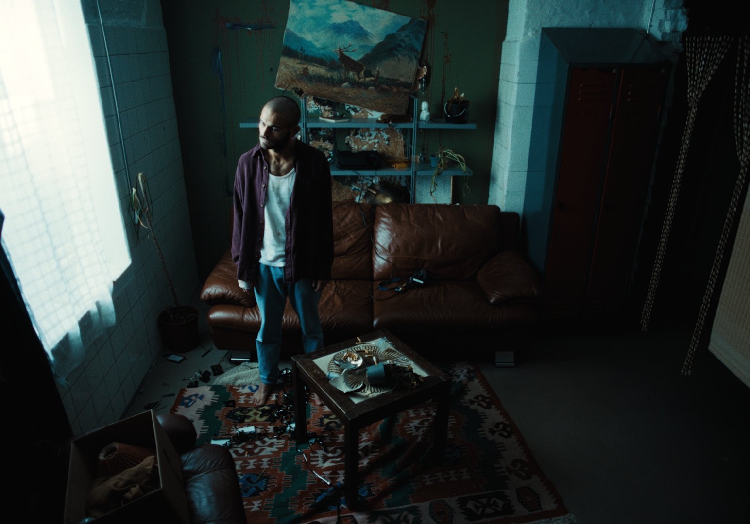 A man standing in a dimly lit living room near a window with blue curtains. The room has a brown leather sofa, a small table with decorative items, and shelves with art supplies or decorations. The floor has a colorful patterned rug, and various obje
