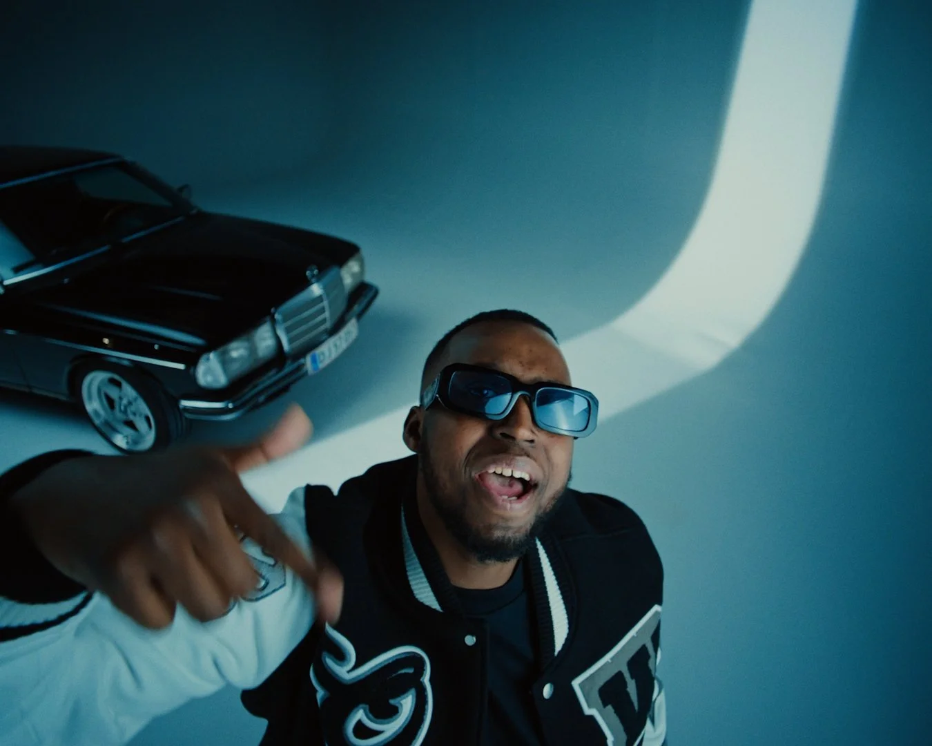 A man with sunglasses and a beard smiling and making a gesture with his hand, with a toy car on the ground beside him against a plain gray background.