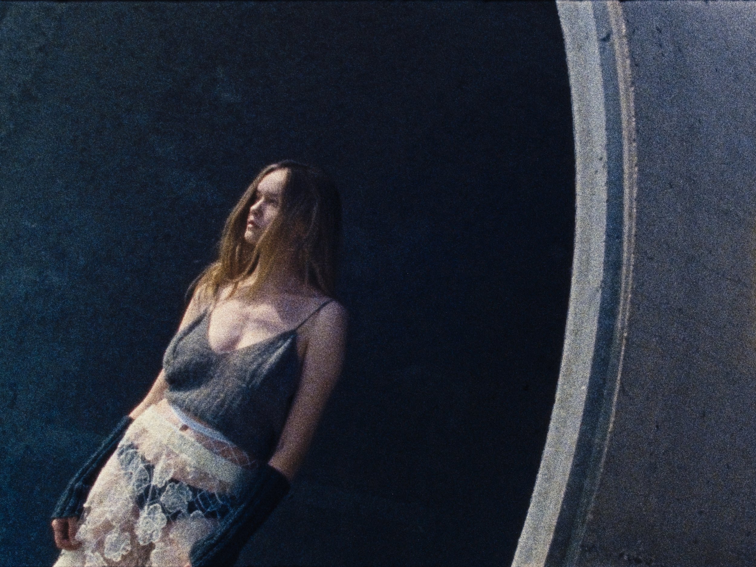 A young woman with long hair leaning against a dark wall, wearing a spaghetti strap top and lace-patterned shorts, with her eyes closed and head tilted slightly.