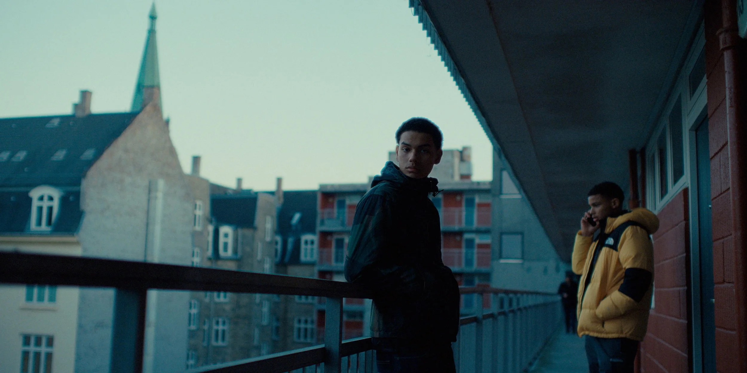 Two young men on a balcony in an urban setting, one in a black jacket looking towards the camera, the other in a yellow jacket on the phone, with buildings and a cloudy sky in the background.