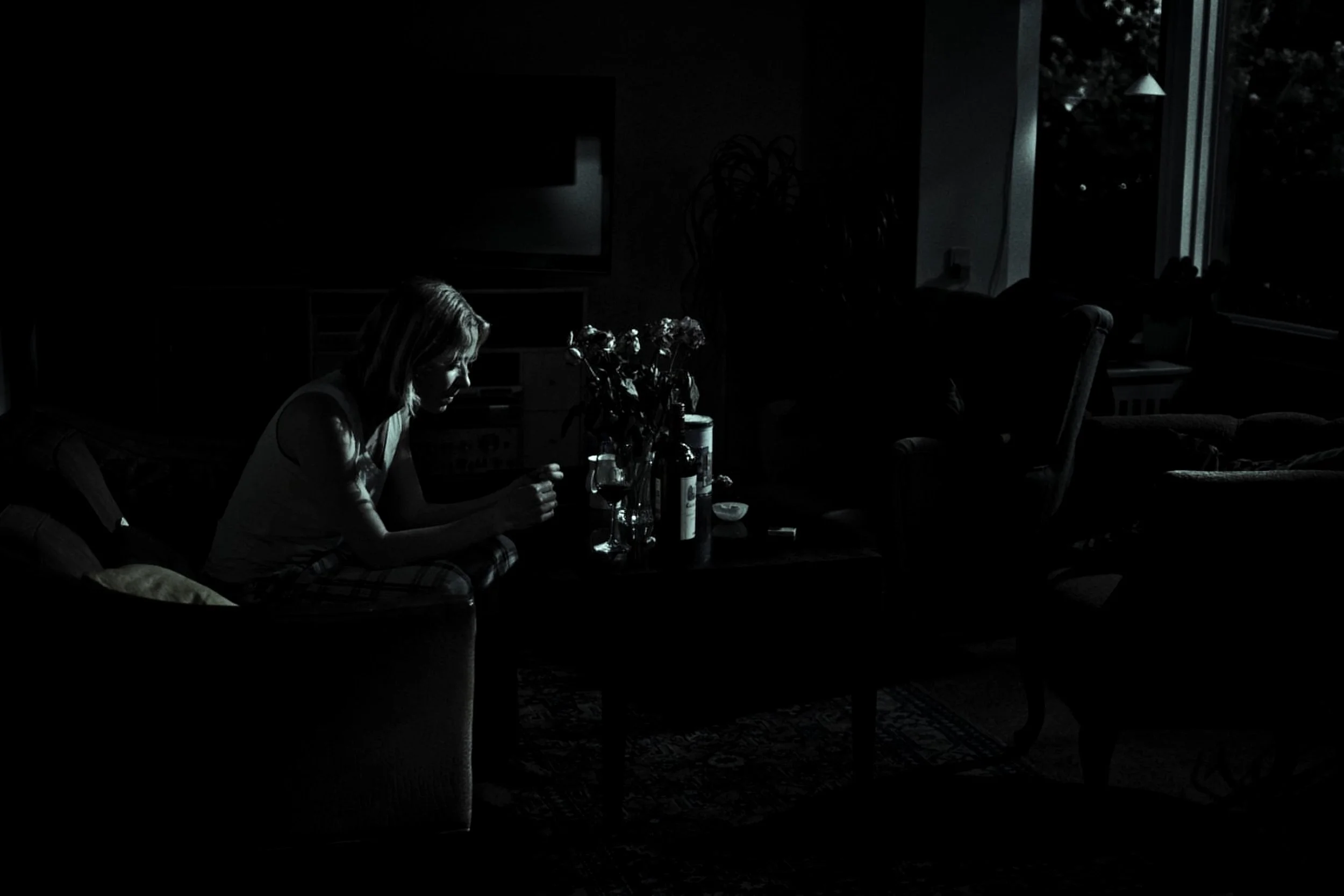 A woman sitting on a sofa in a dark living room, illuminated by natural light coming through windows, with a table in front of her holding flowers, bottles, and a wine glass.