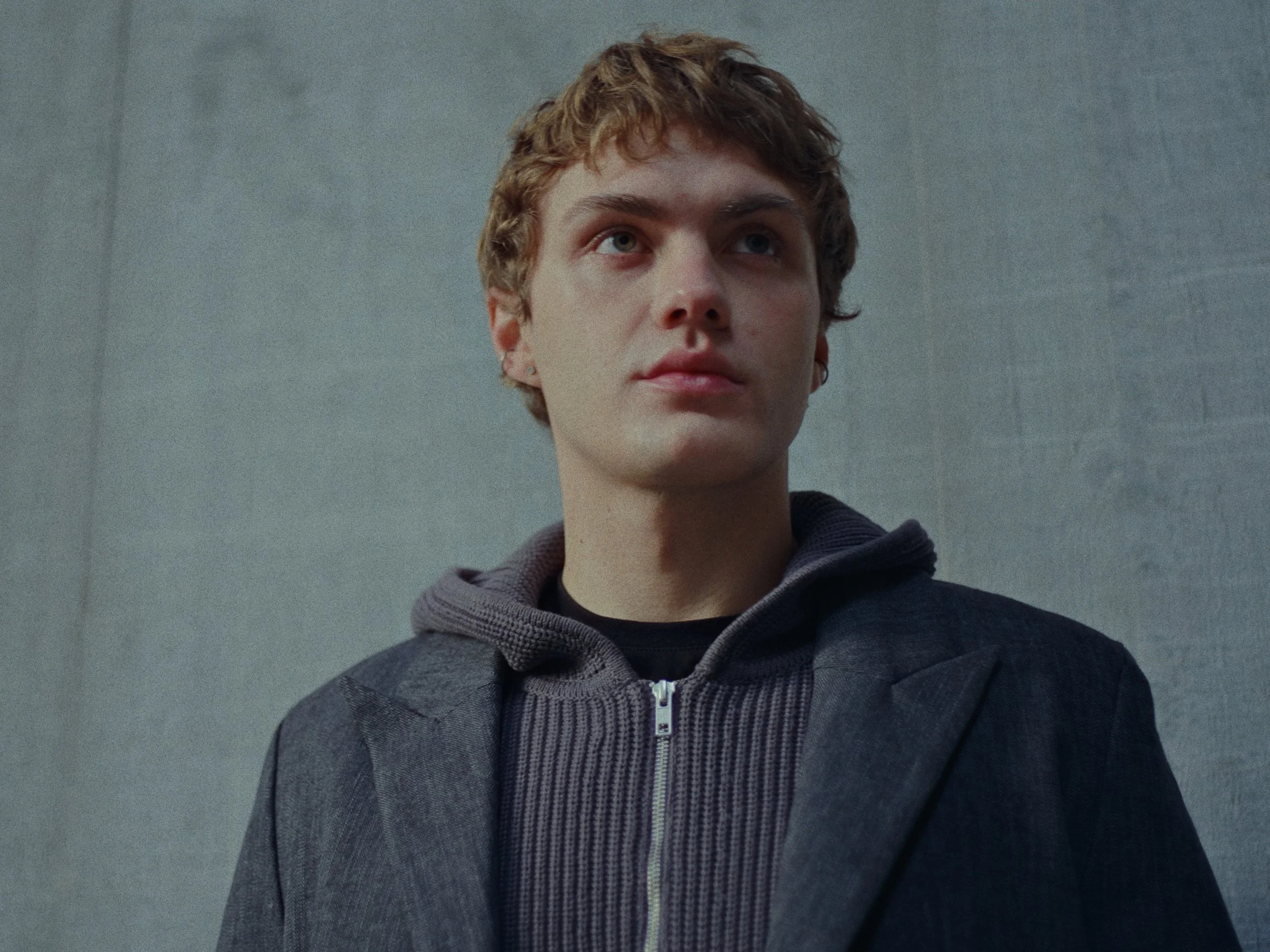 A young man with curly blonde hair, wearing a dark blazer over a hoodie, standing against a gray wall.