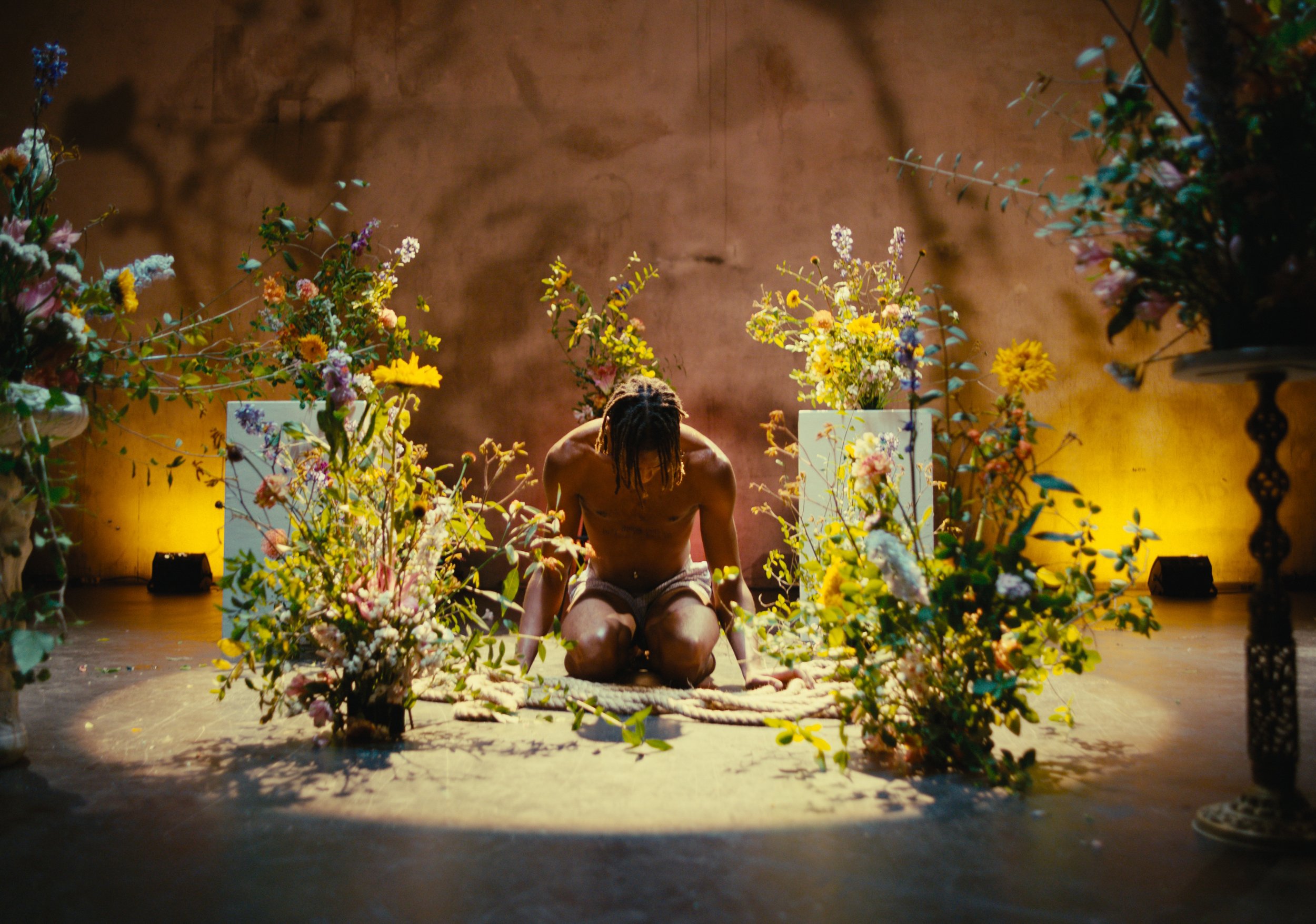 A man kneeling on the floor surrounded by colorful flowers in a dimly lit room with warm yellow lighting.