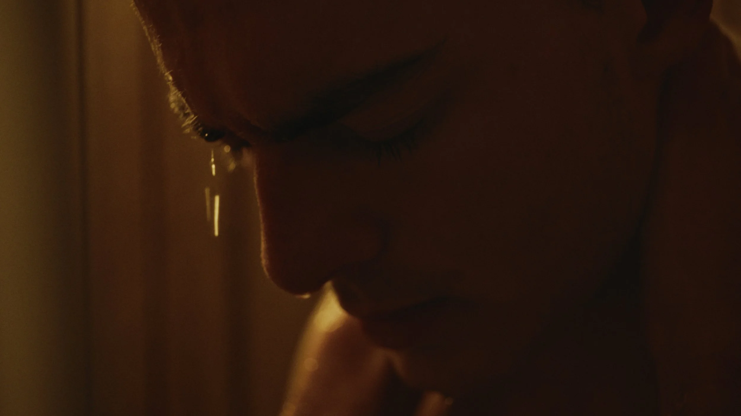 Close-up of a person's face with closed eyes, with a tear falling down the cheek in warm, subdued lighting.
