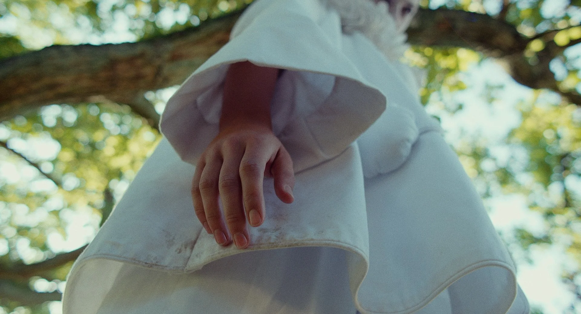 Close-up of a person's hand resting on their thigh while sitting on a tree branch, with green leaves and sunlight in the background.