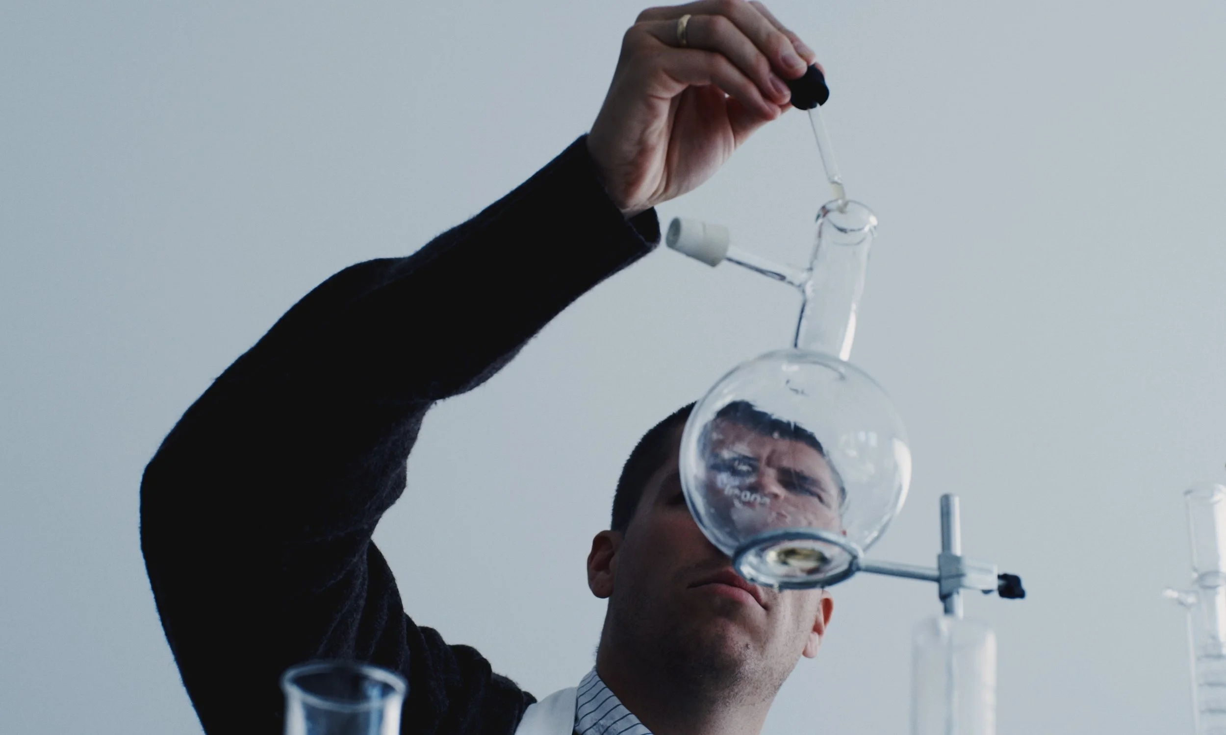 A man in a dark sweater and dress shirt performs an experiment in a laboratory, pouring liquid from a glass container into another held above his head.