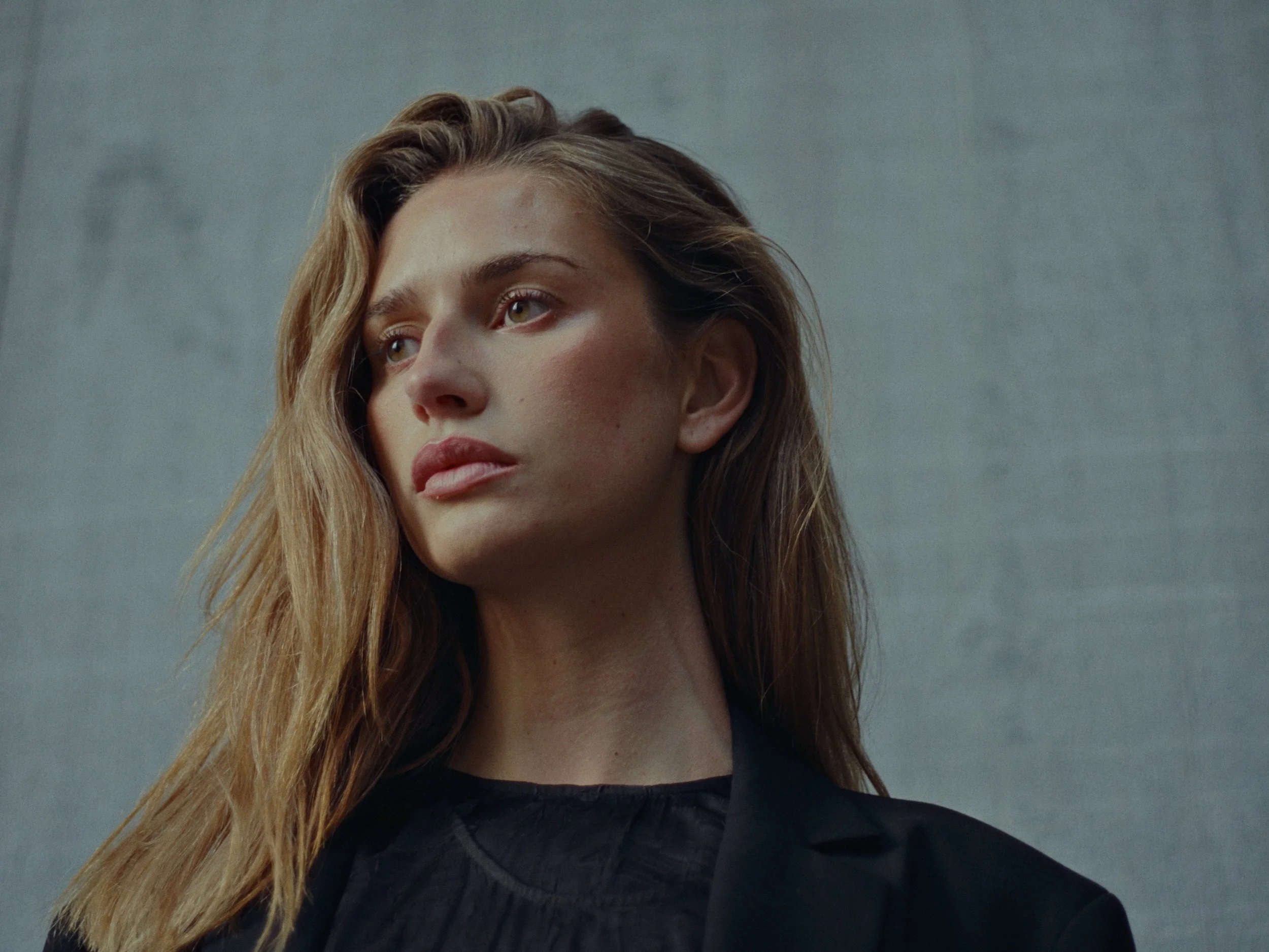 Close-up of a young woman with long, wavy light brown hair, looking thoughtfully to the side against a plain gray background.