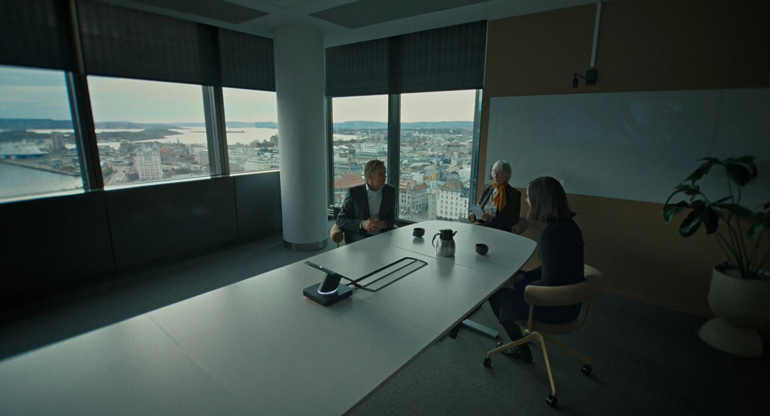 Three people in a conference room having a discussion with cityscape view through large windows.