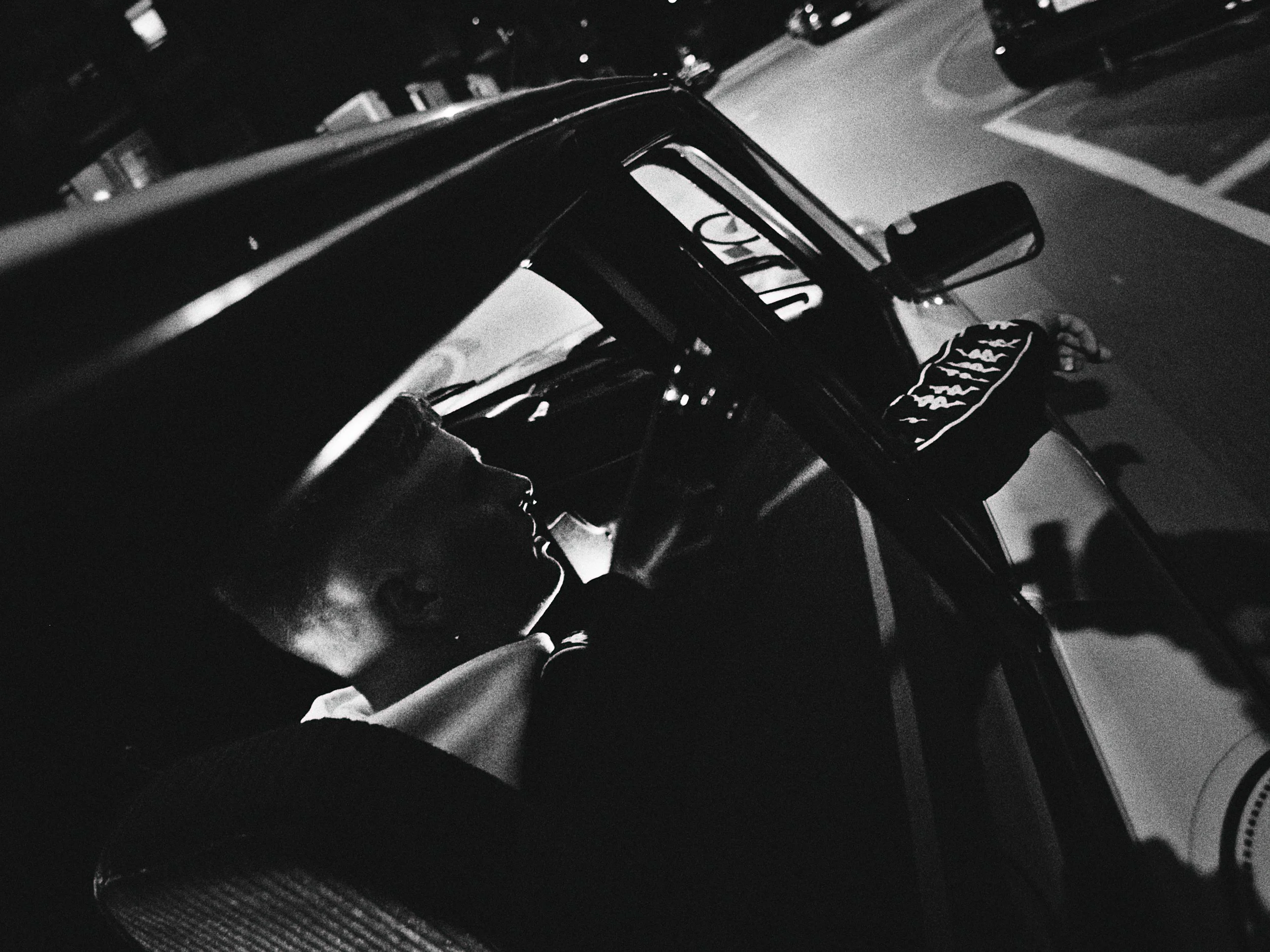 Black and white photo of a person sleeping in the driver's seat of a car at night, with the head resting against the window and the side mirror visible.