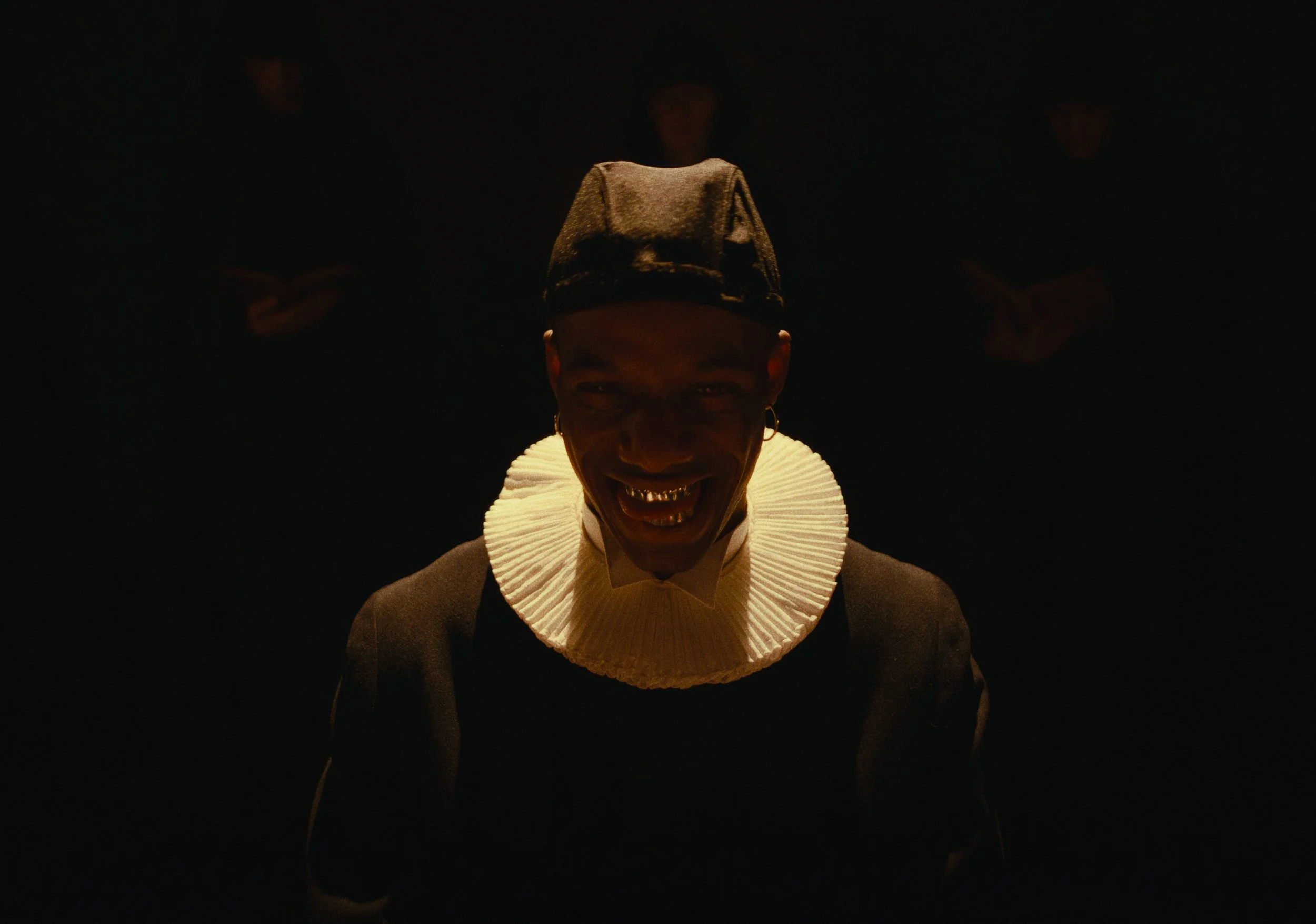 A person with dark skin smiling or sneering, wearing a hat, gold teeth, hoop earrings, and a ruffled white collar, in a dark setting with a spotlight on their face.