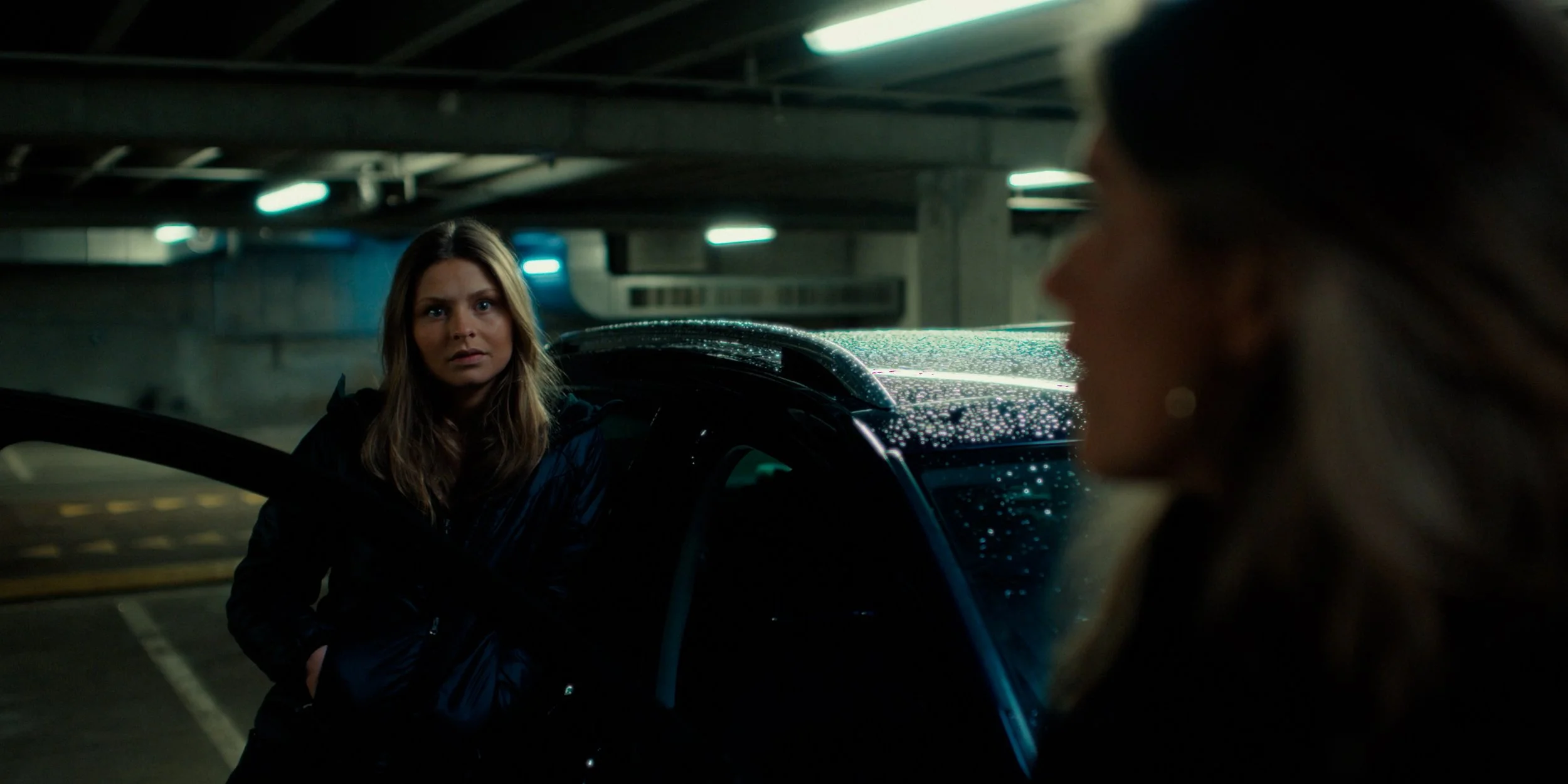 Two women are having a conversation in a dimly lit, underground parking garage, with one woman facing the camera and the other with her profile visible.