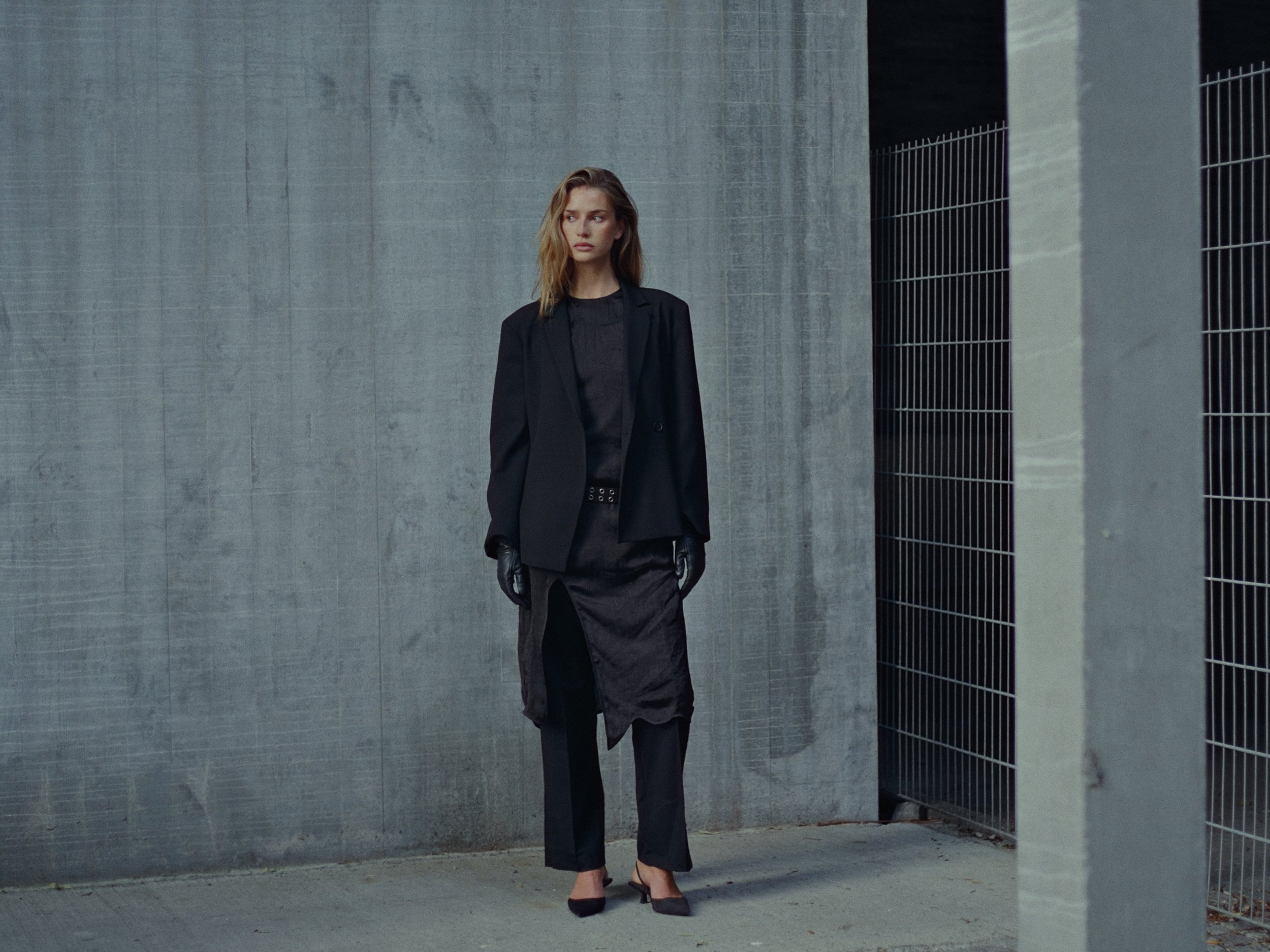 A young woman standing against a concrete wall in an urban setting, dressed in black fashion, wearing a blazer, skirt, and gloves, with a serious expression.