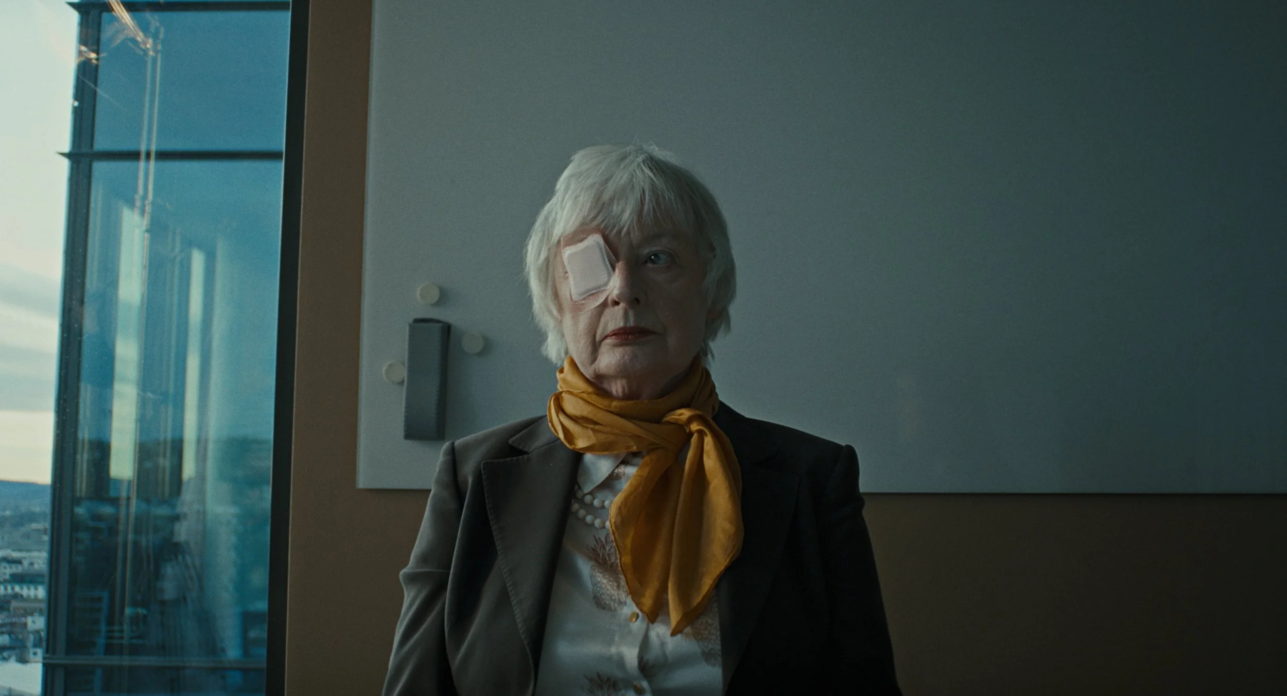 An elderly woman with white hair and an eye patch over her left eye, wearing a black blazer and a mustard yellow scarf, standing indoors near a large window with a view of a cityscape at sunset.