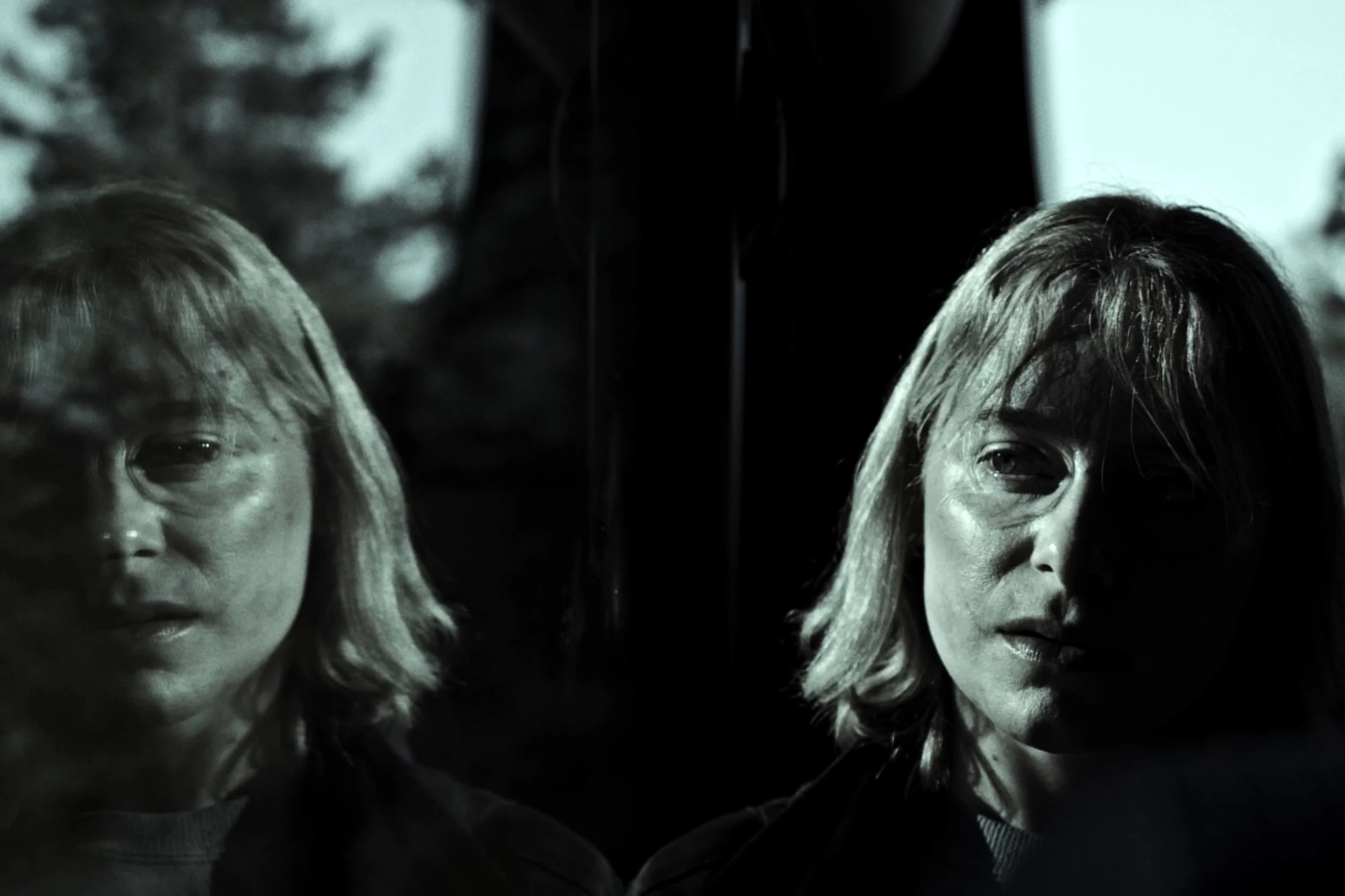 A black and white photo of a woman’s face reflected in a glass window. The woman's face appears on both sides of the glass, creating a symmetrical effect.