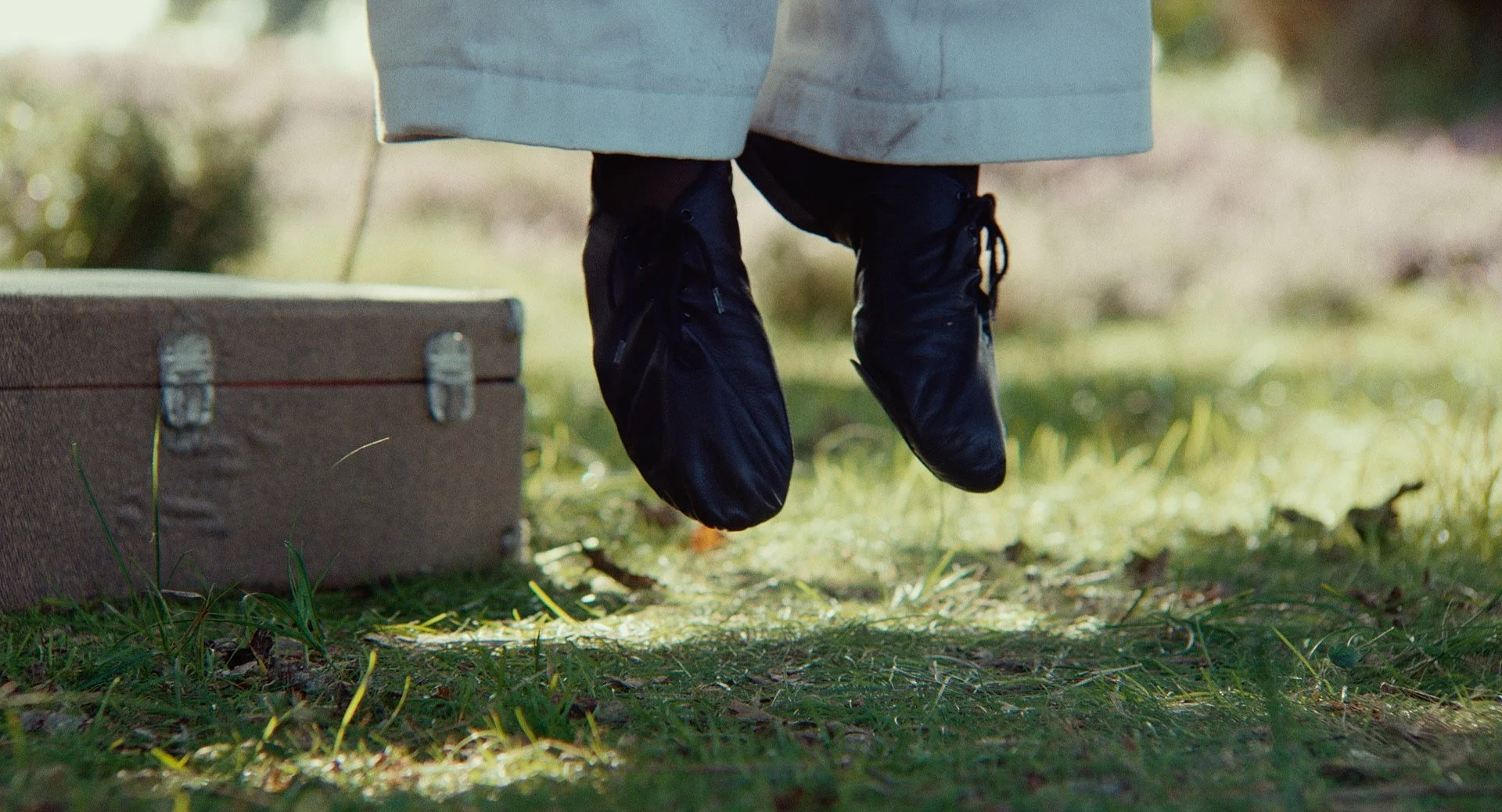 A pair of black shoes hanging from a person's feet, with the person standing on grass near a brown wooden box.