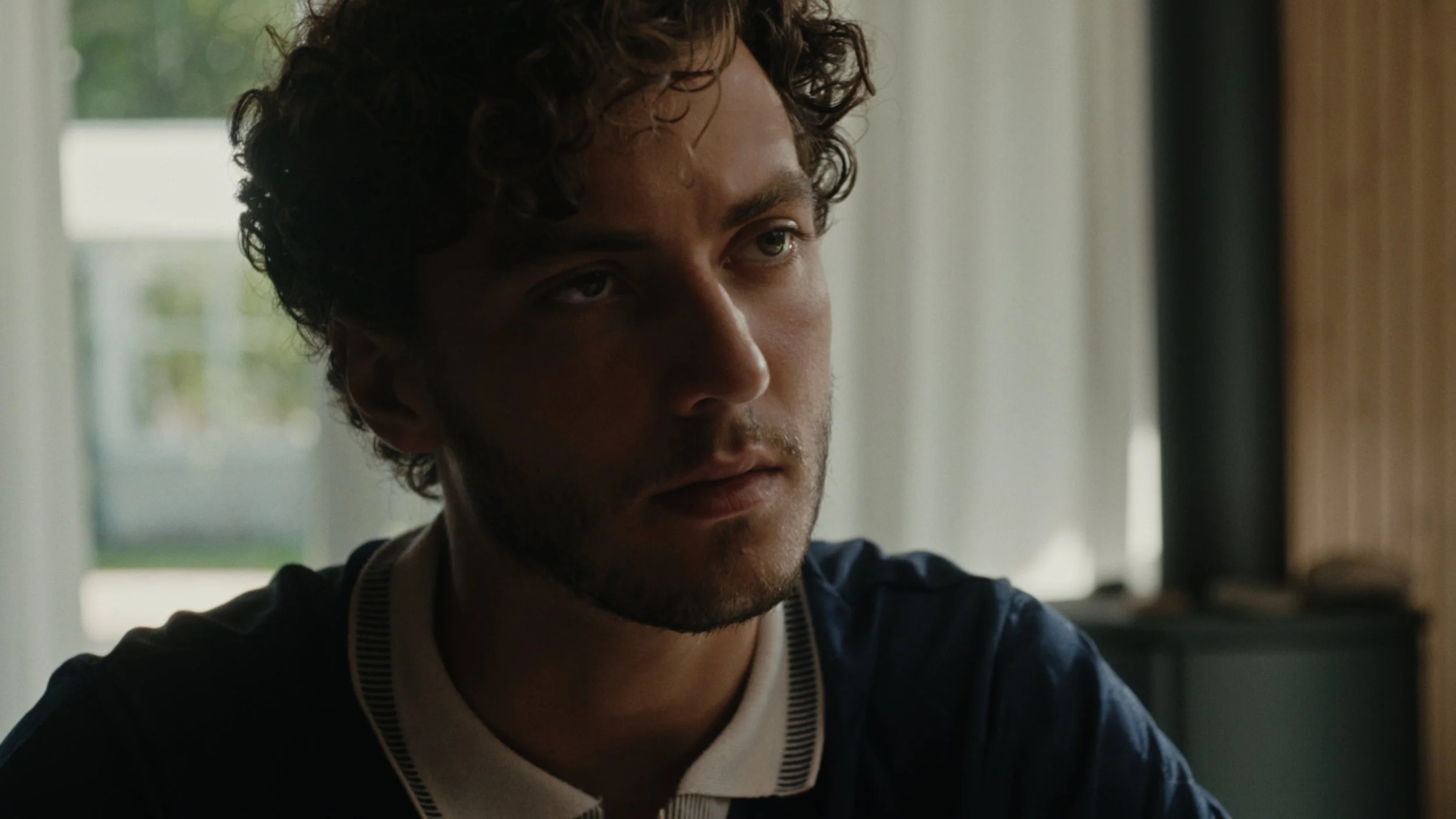 A young man with curly hair and facial hair sits indoors near a window, looking to the side with a serious expression.