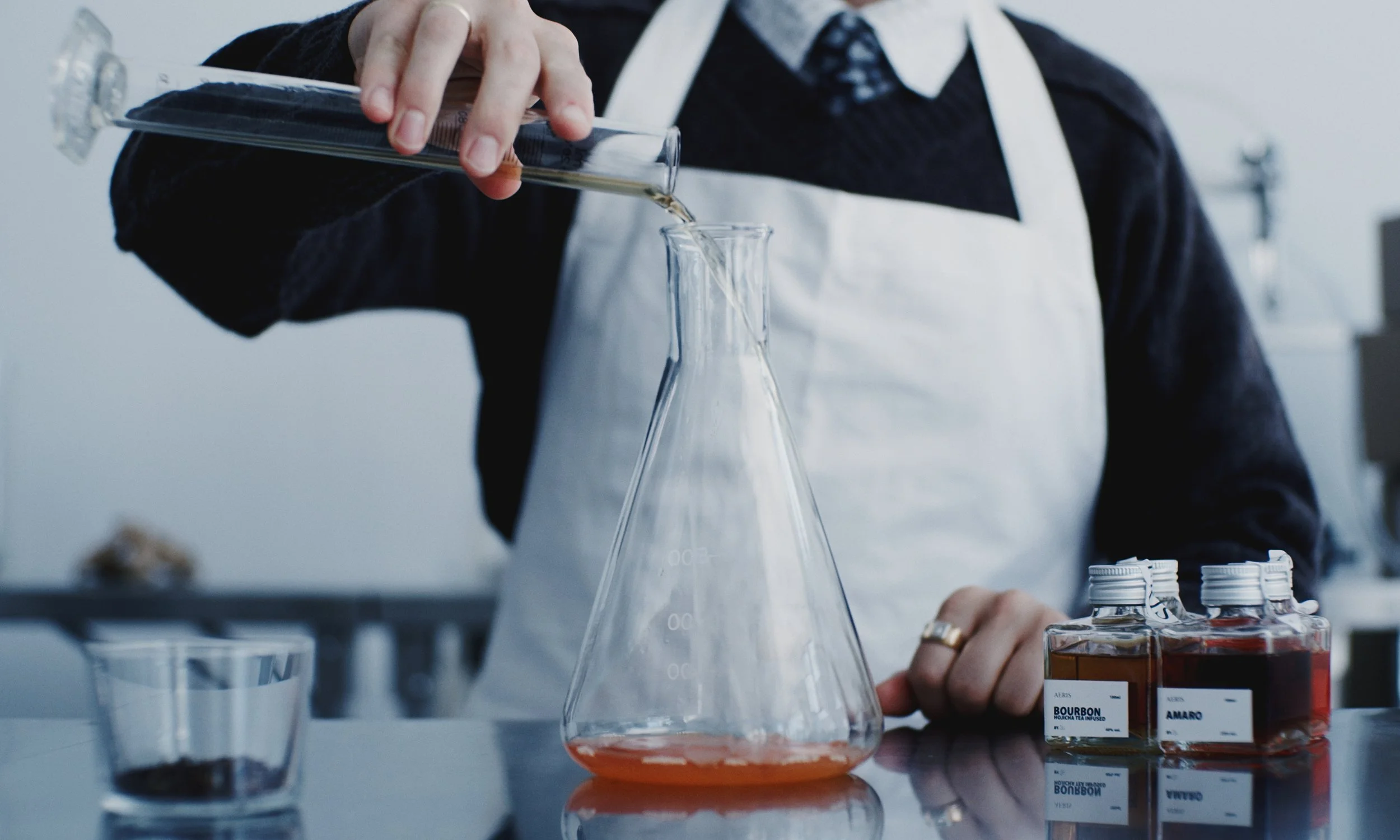 Person in a lab coat and apron pouring a liquid into a glass flask in a laboratory setting.
