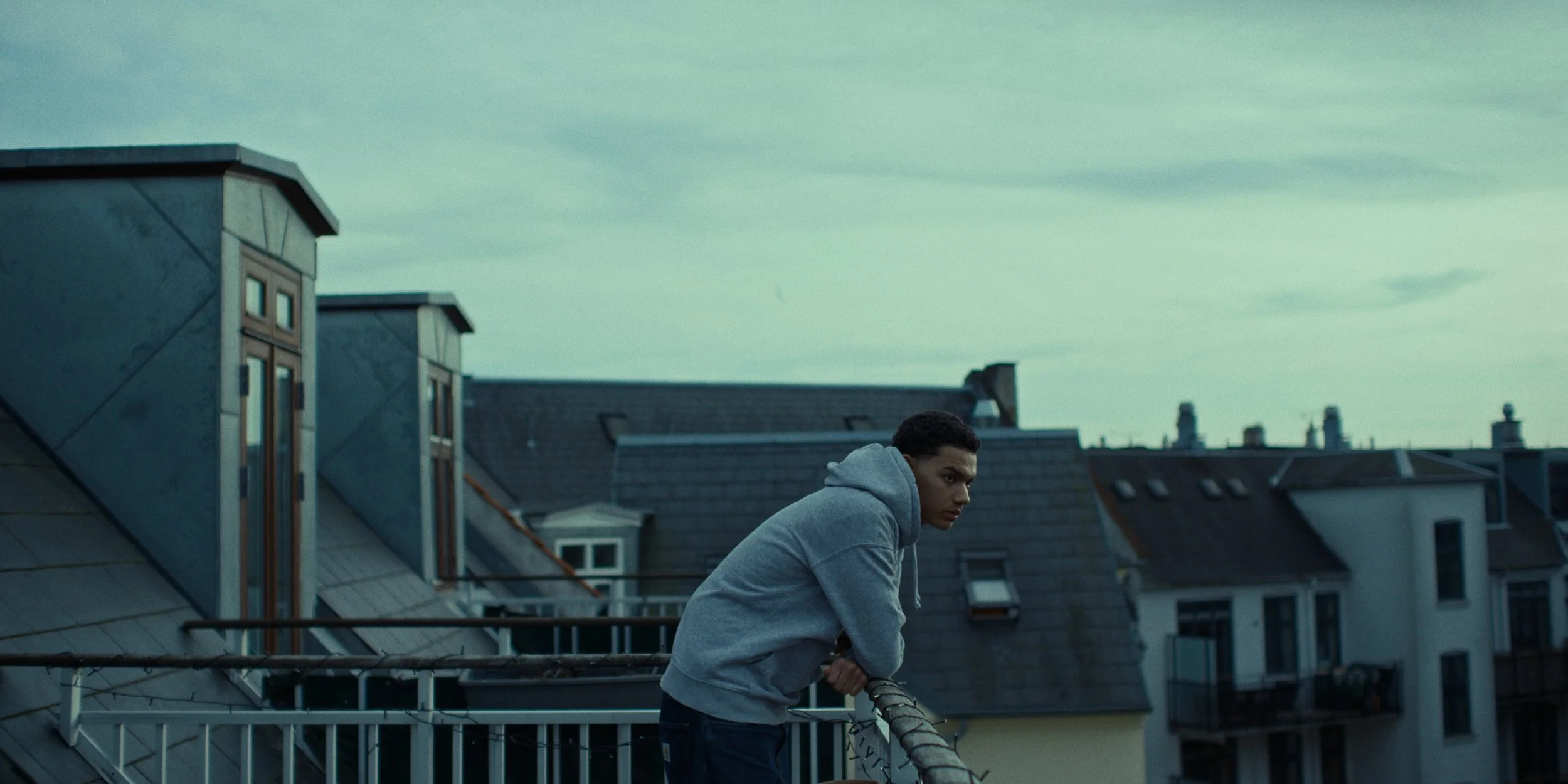A young man wearing a gray hoodie leaning on a balcony railing on a rooftop, with neighboring buildings and an overcast sky in the background.