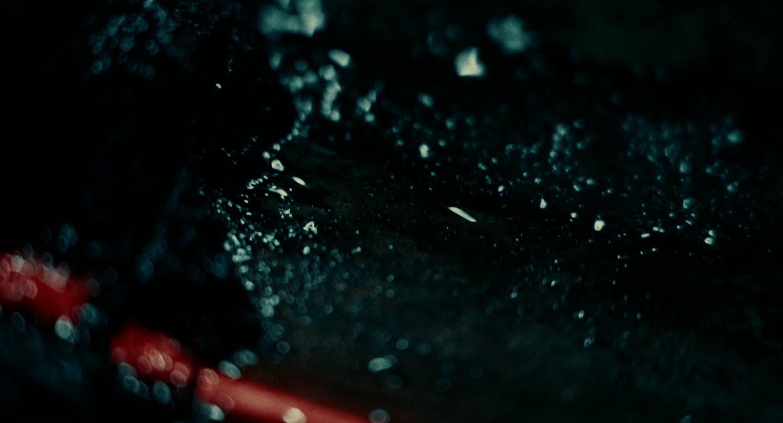Close-up of a dark, wet surface with small, glistening water droplets and a red object partially visible in the lower left corner.