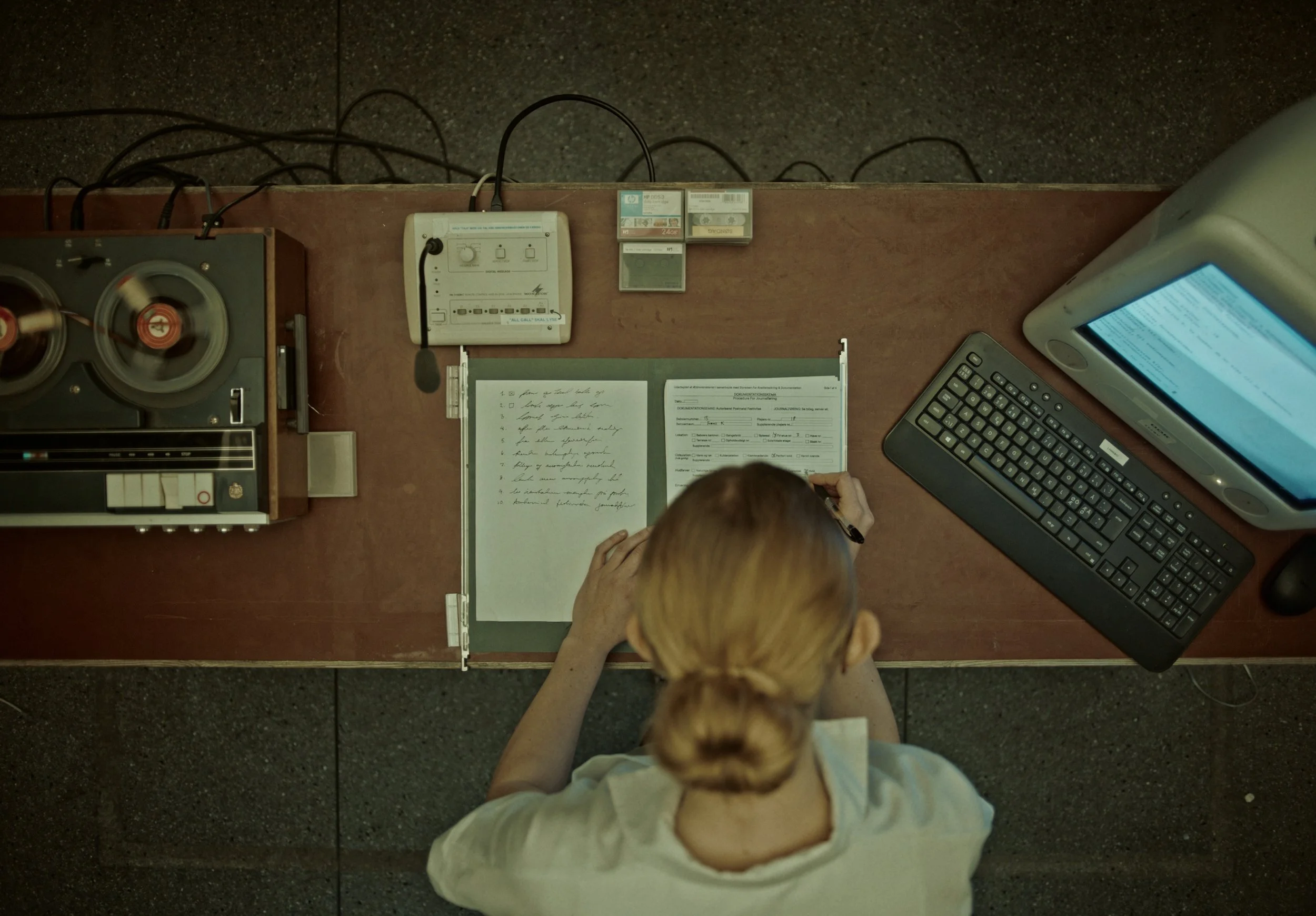 Woman with blond hair in a bun working at a desk with a notebook, vintage tape recorder, typewriter, monitor, keyboard, and various office supplies.