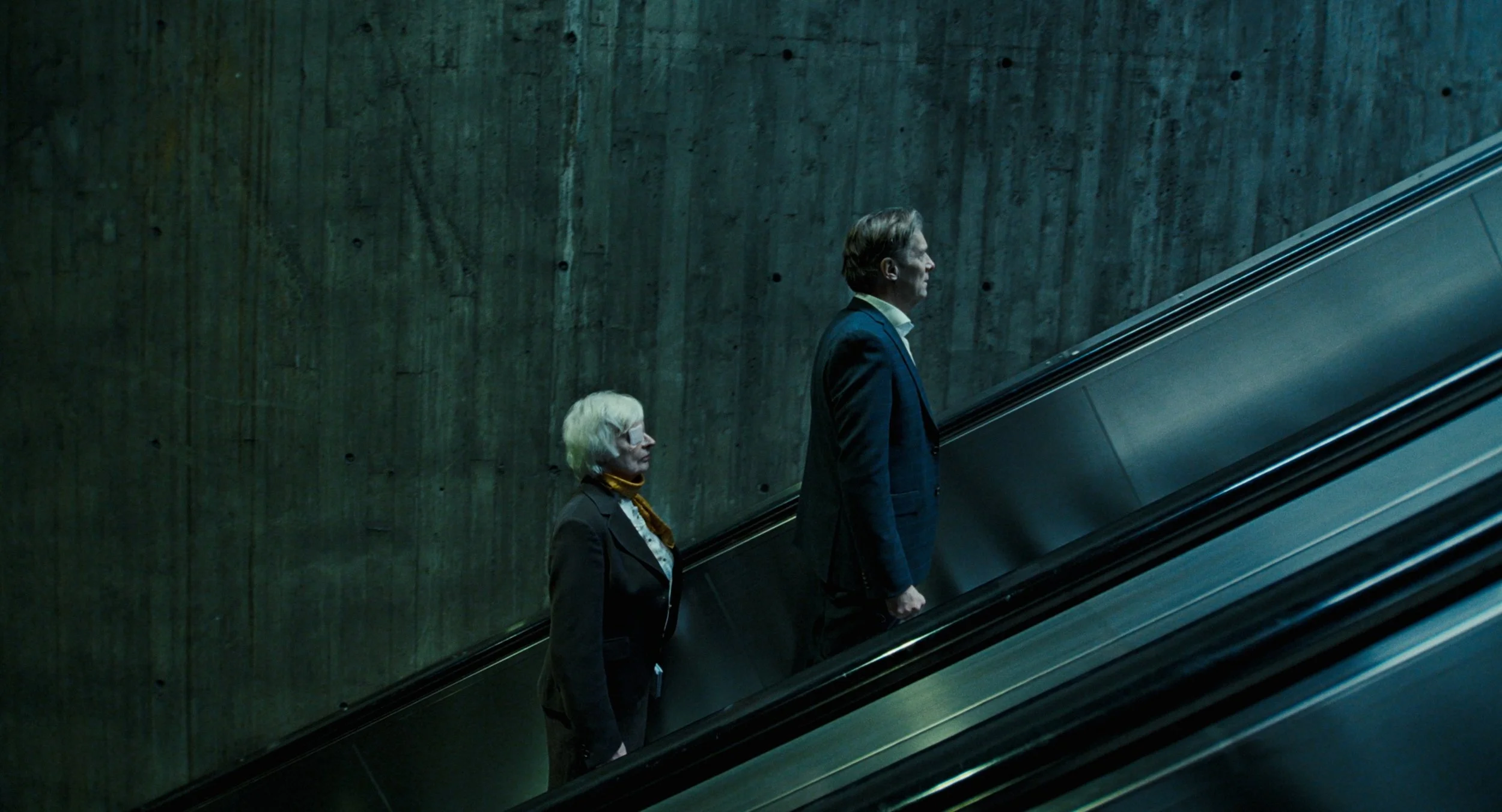 A man and a woman in business attire riding an escalator in a dimly lit, concrete-walled setting.
