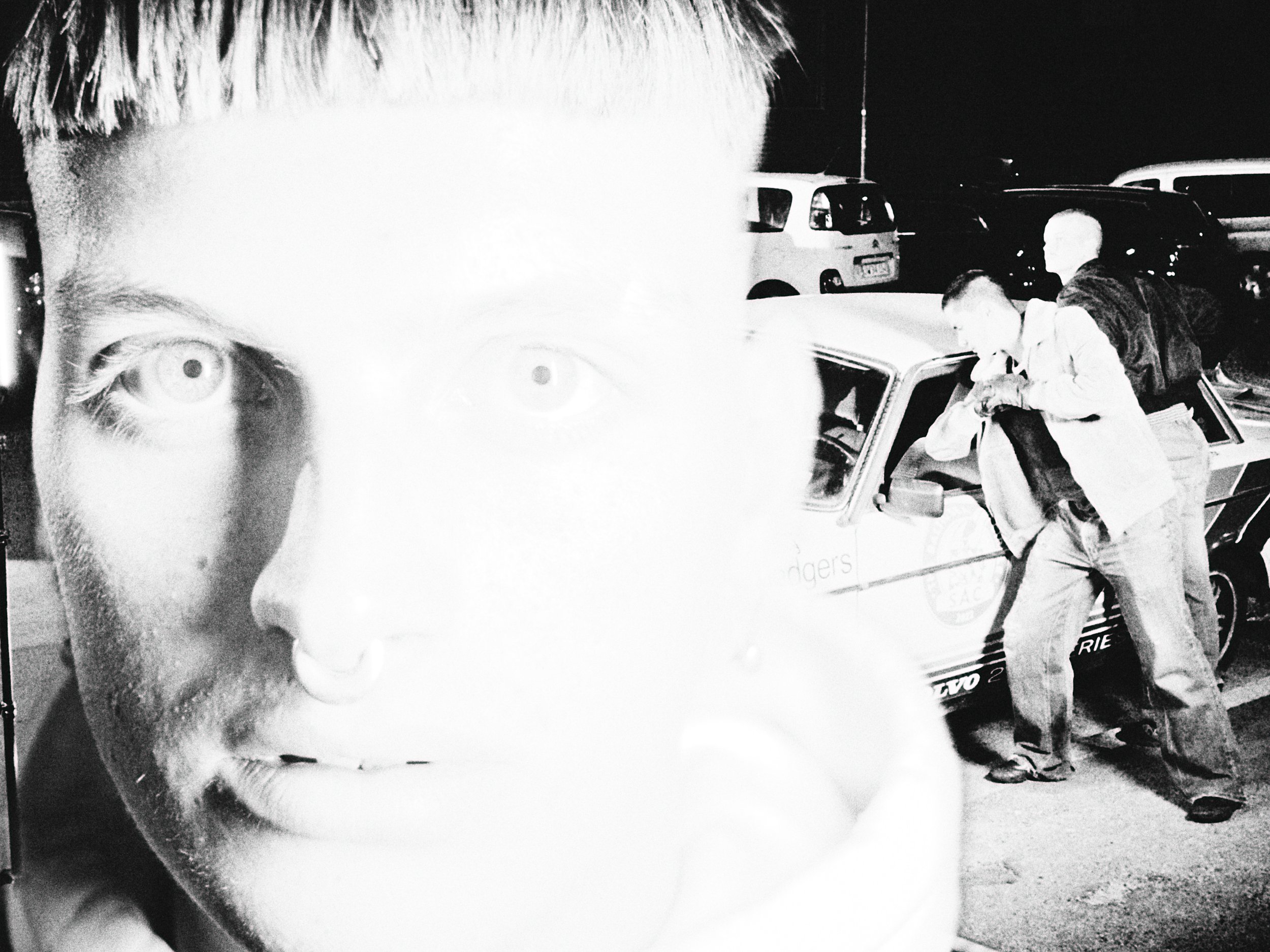 Black and white photo of a woman holding a poster of a young man's face, with police officers arresting another person near a car in the background at night.