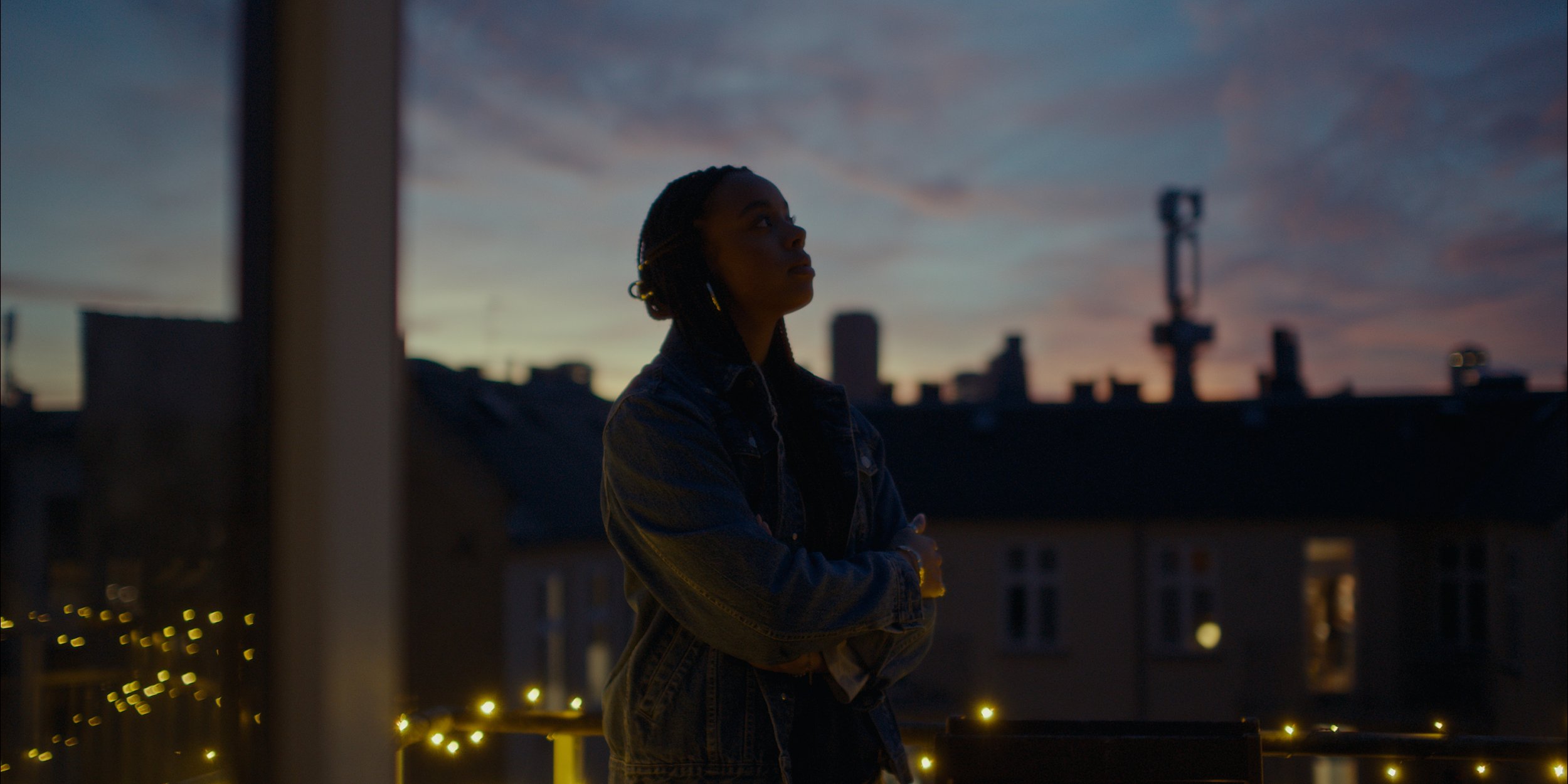A woman stands on a balcony during sunset, gazing into the distance with her arms crossed. The city skyline and buildings are visible in the background, with some windows lit and string lights illuminating the balcony.