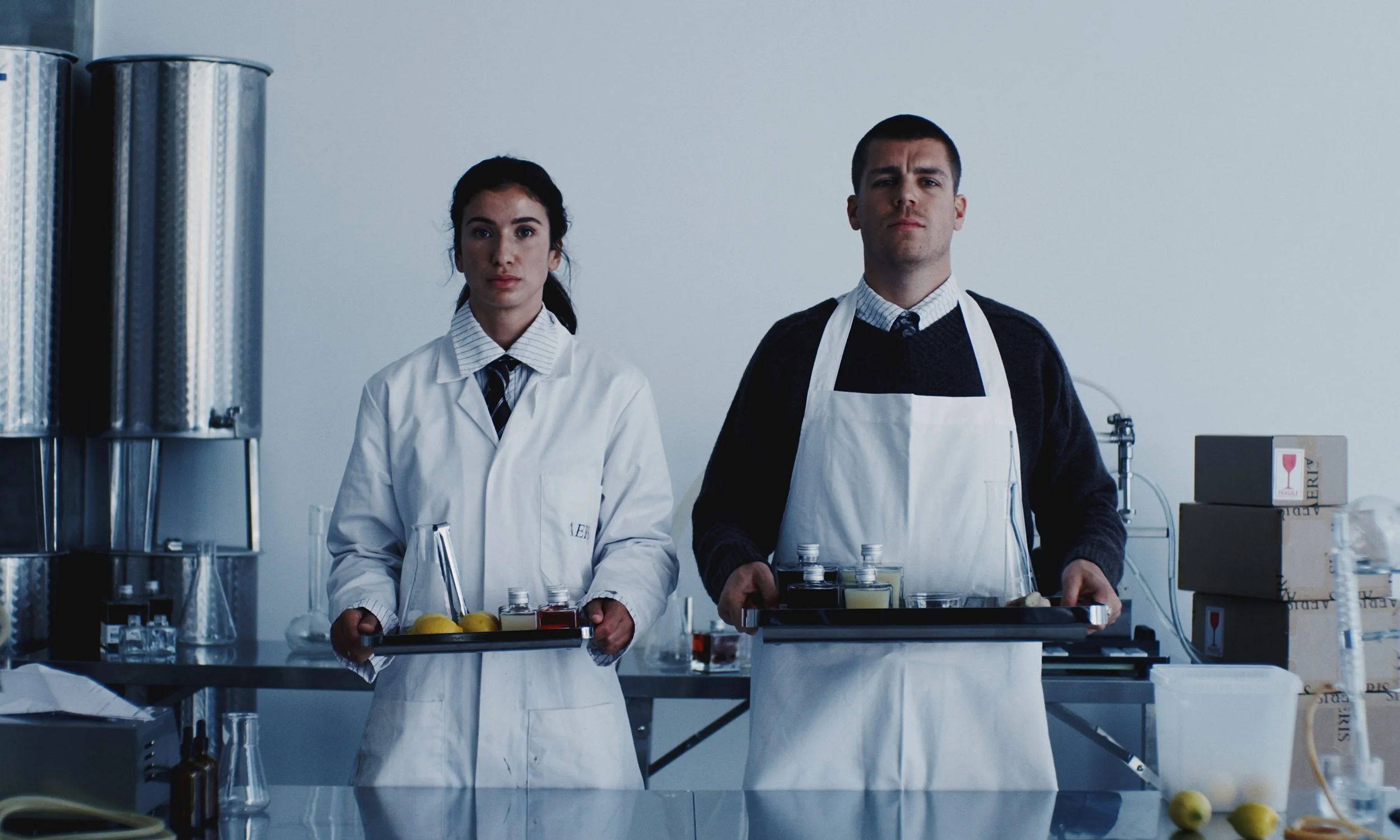 Two bartenders in a laboratory-like setting, holding trays with bottles of ingredients, lemon, and equipment, wearing white aprons and standing behind a stainless steel counter.