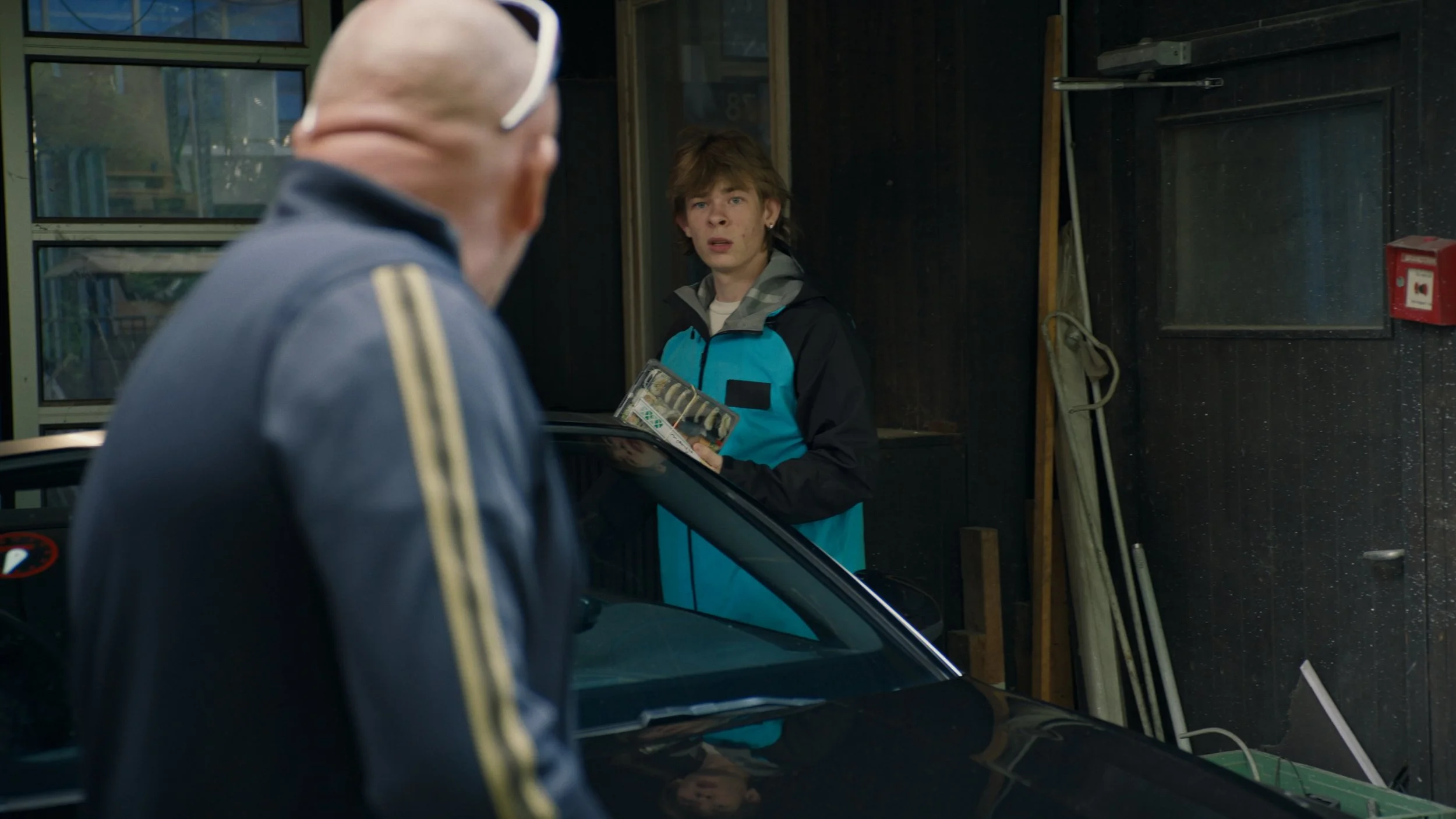A young man in a blue and black jacket holding a magazine, looking surprised, standing inside a garage. An older man with a bald head and sunglasses on his head, in a dark jacket with gold stripes, is in the foreground with his back to the camera.