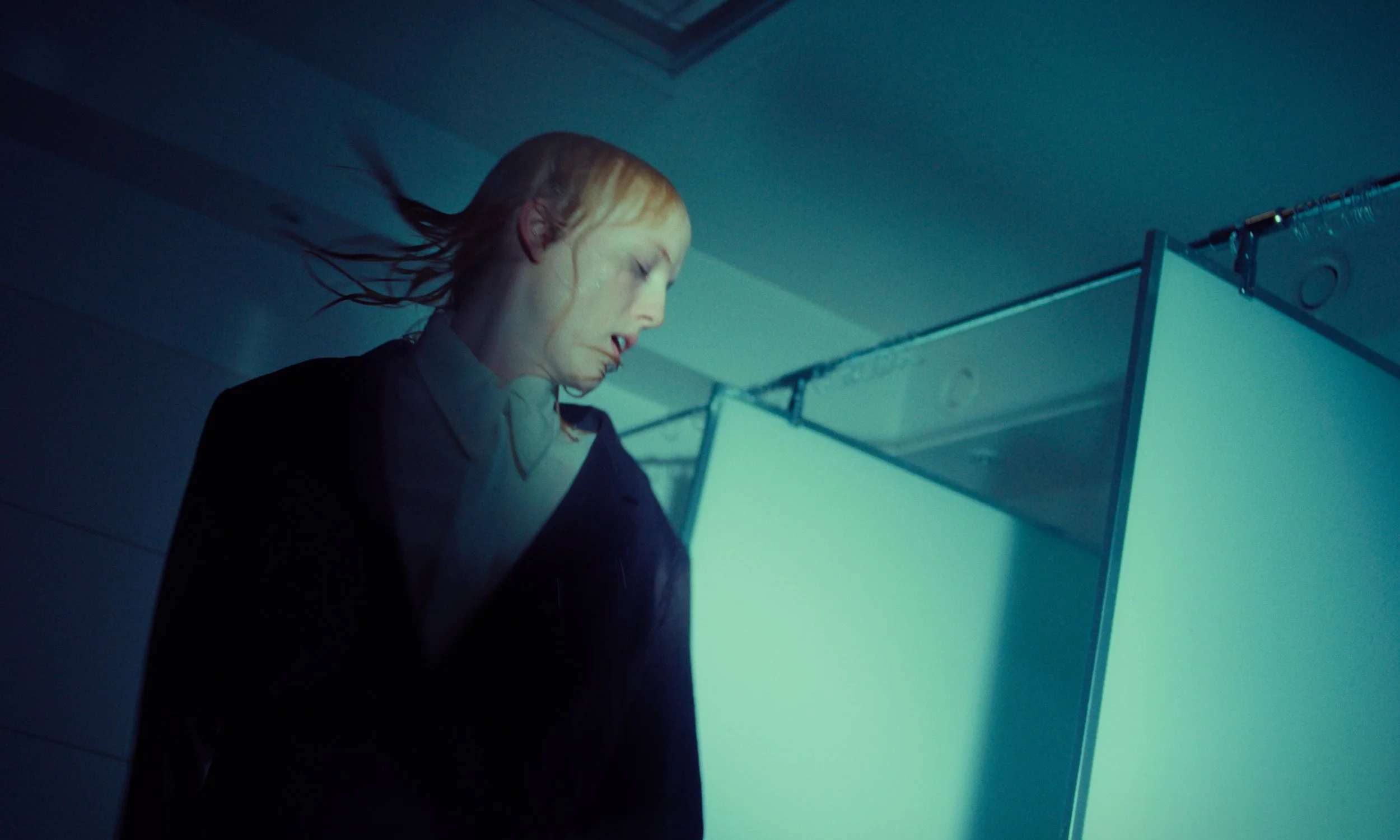 A woman with blonde hair and a white collar stands in a dimly lit room, looking down with her eyes closed, near a light blue partition or divider.