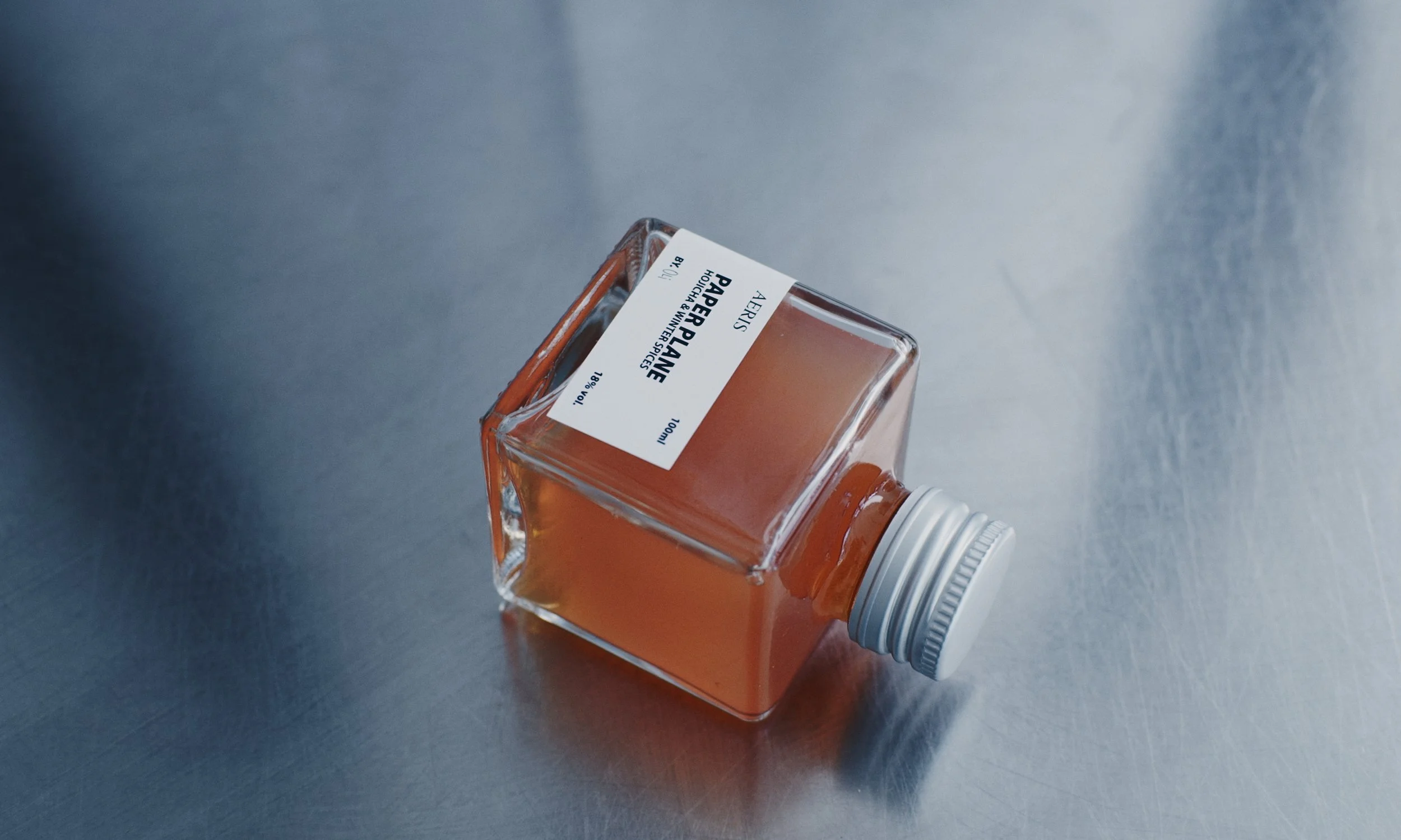 A square glass bottle lying on its side, containing a pinkish liquid, with a white screw cap and a white label that reads "Arbes Paper Palm".