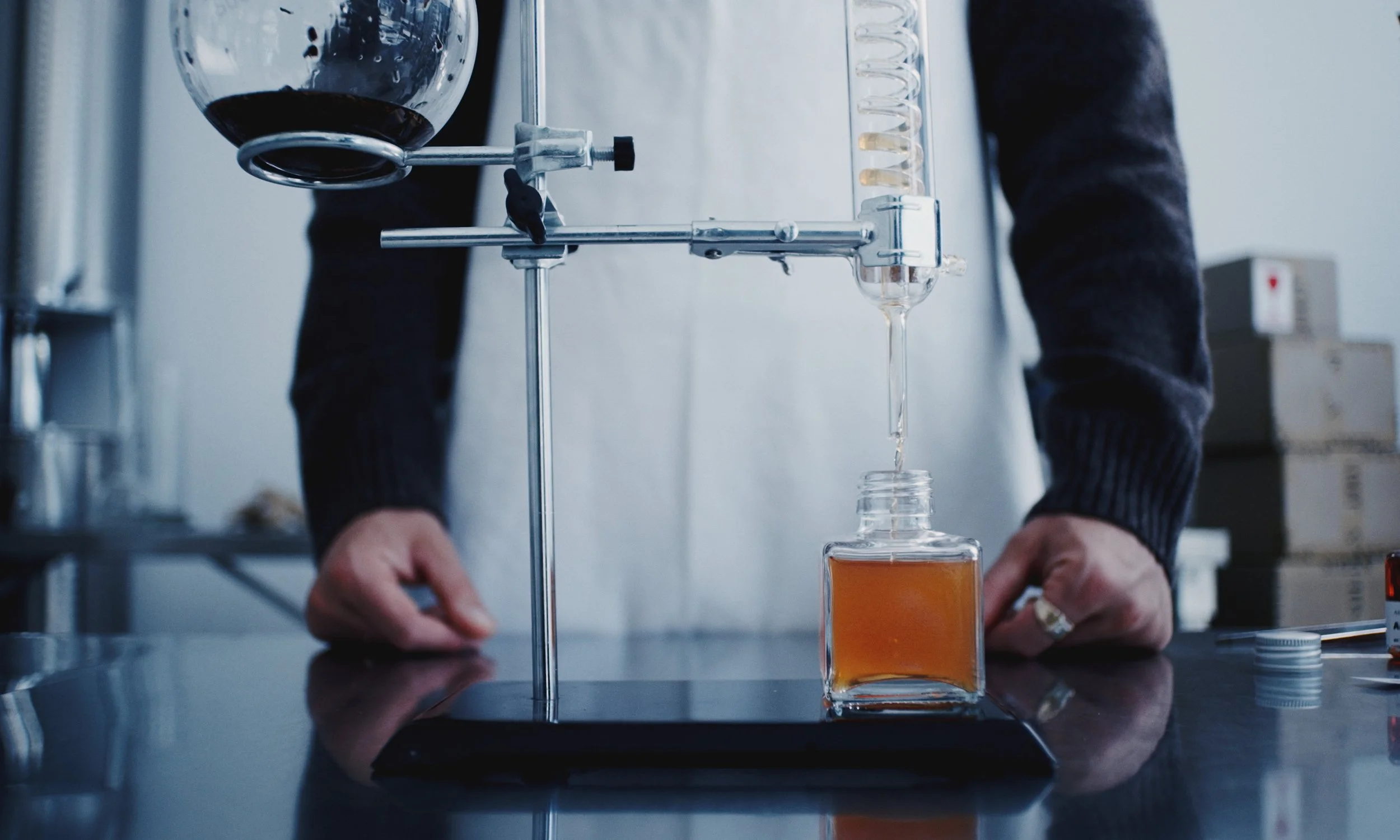 A person in a lab coat conducting a chemistry experiment with glassware and liquids on a black surface.