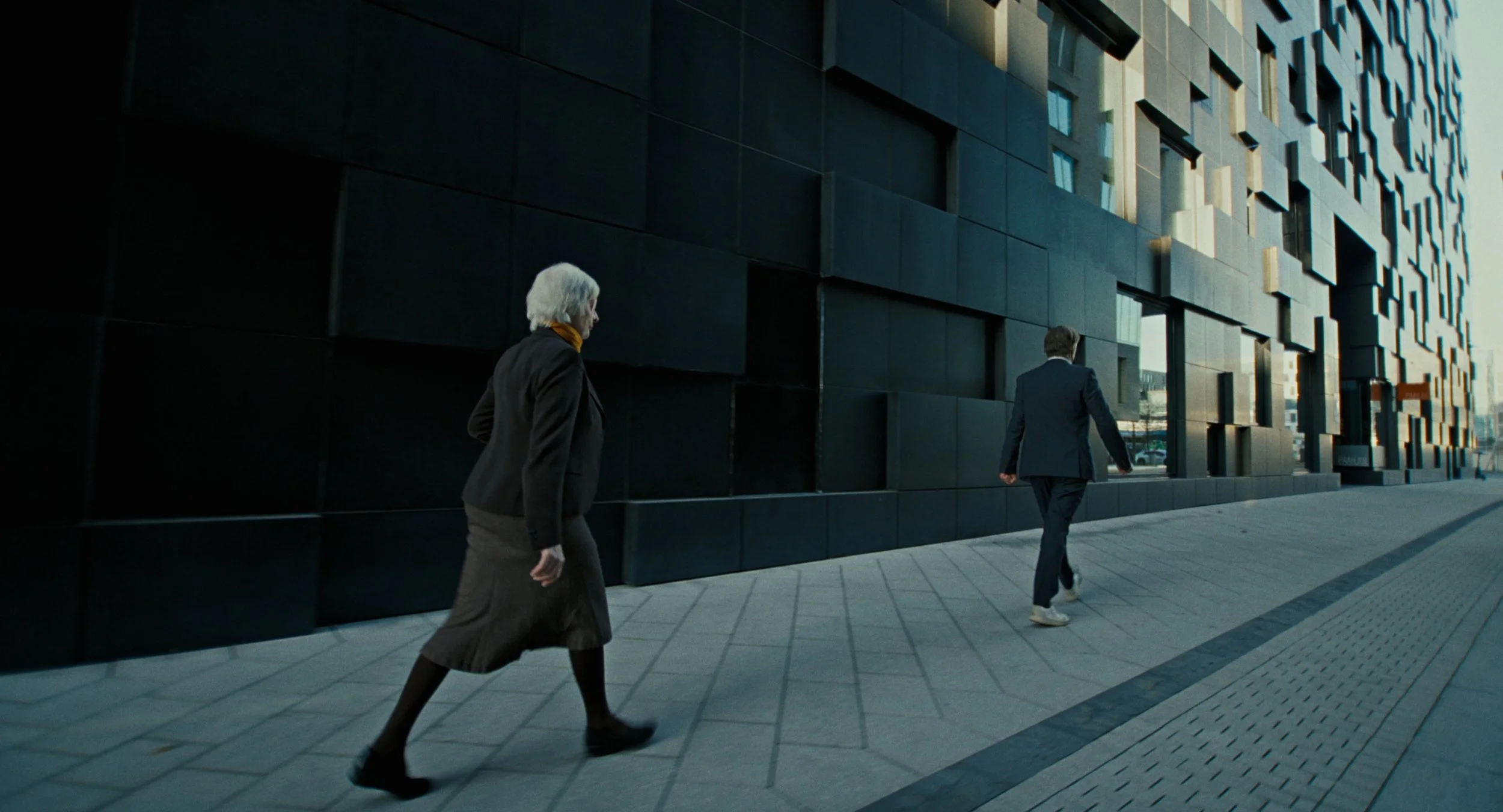 Two people walking along a sidewalk in front of a modern, dark-colored building with textured square panels.