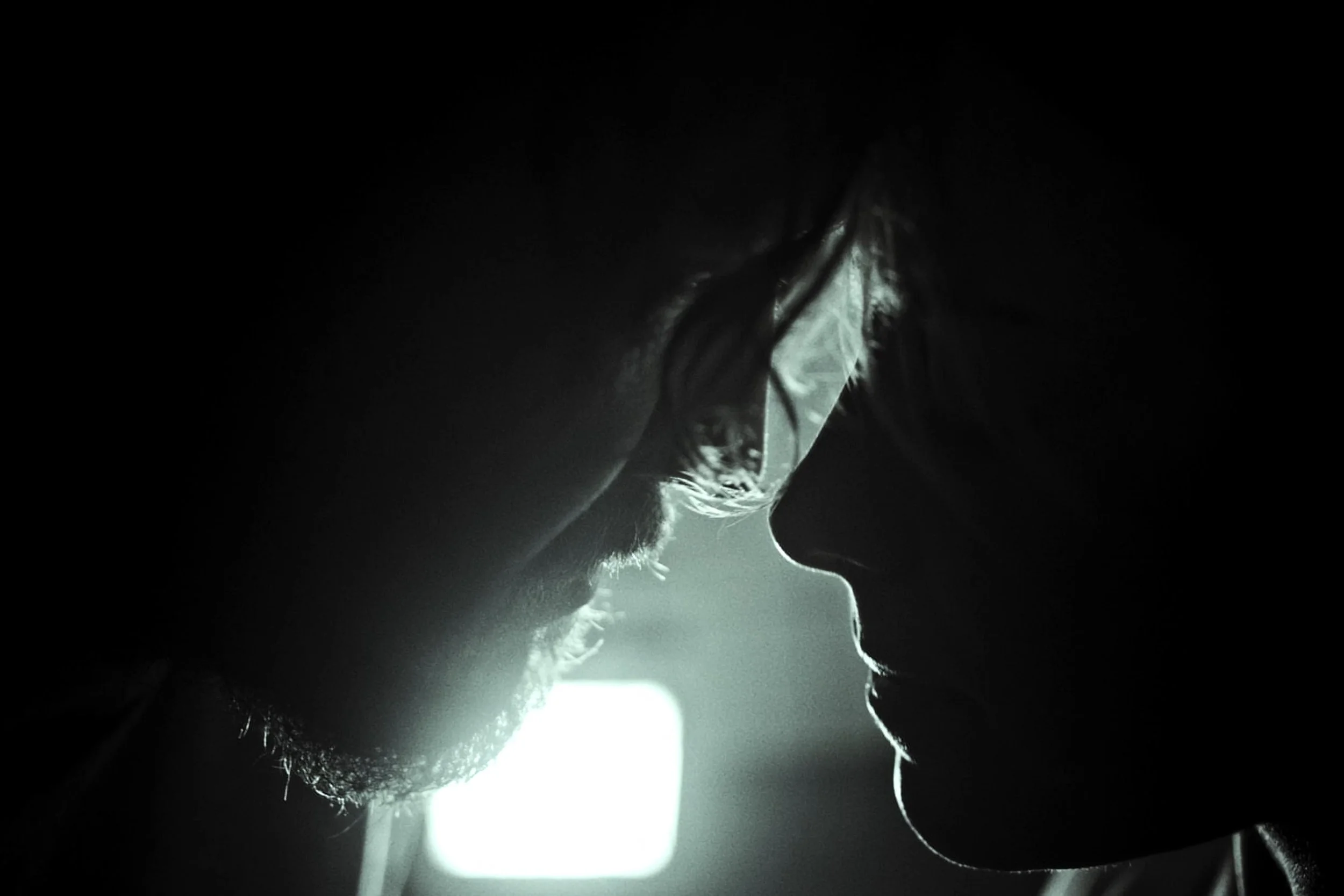 Close-up black and white photograph of two people facing each other closely, in silhouette, with a soft light behind them highlighting their profiles.