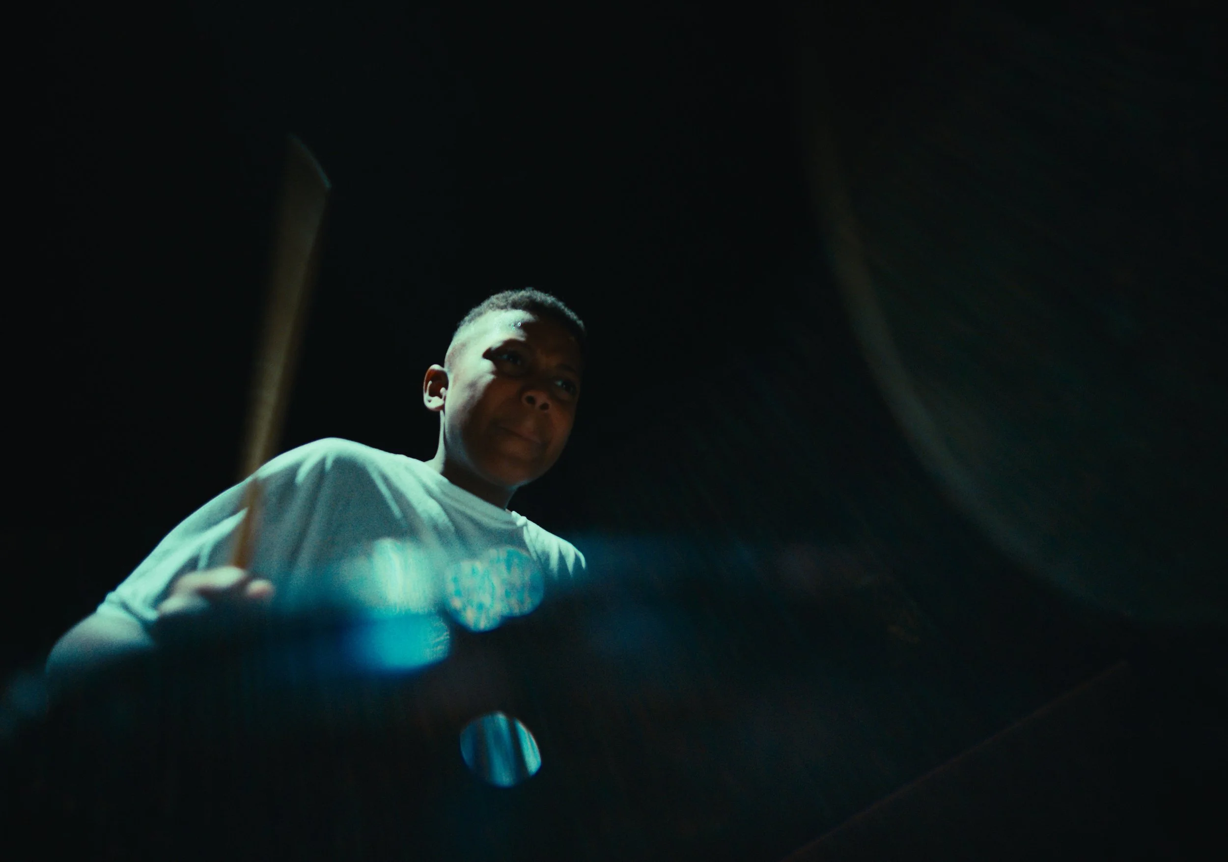 A young boy is playing a drum kit in a dimly lit room, with a focused expression on his face.