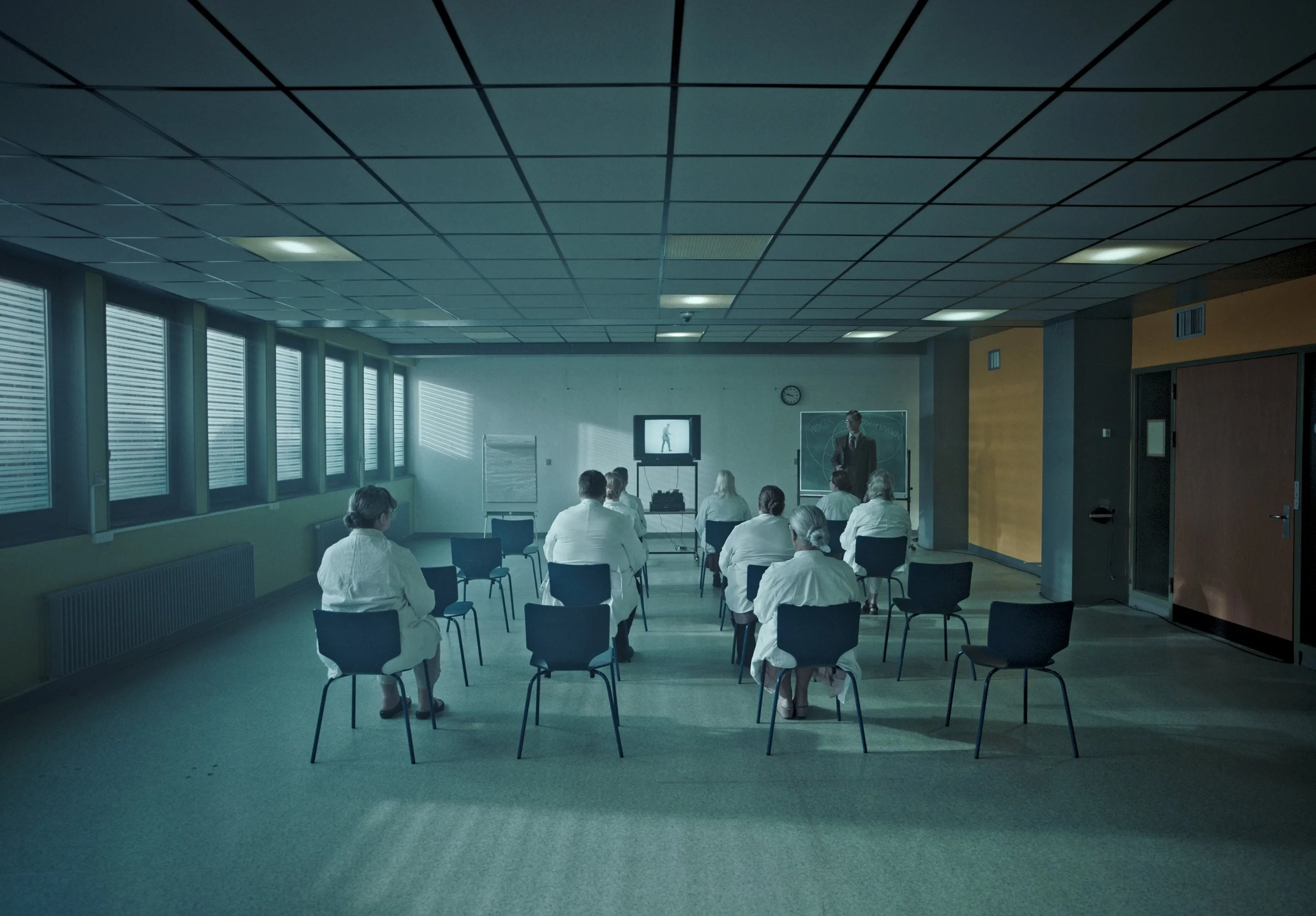 A group of people in hospital gowns attending a presentation in a large, well-lit room with green walls and a ceiling with square tiles. The presenter is at the front, near a whiteboard and chalkboard, with a TV screen showing a person walking. The r