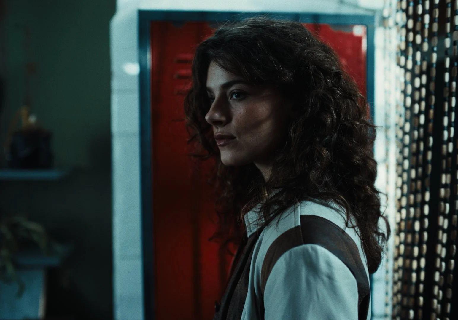 Side profile of a woman with curly hair standing in front of a red locker and a patterned curtain.