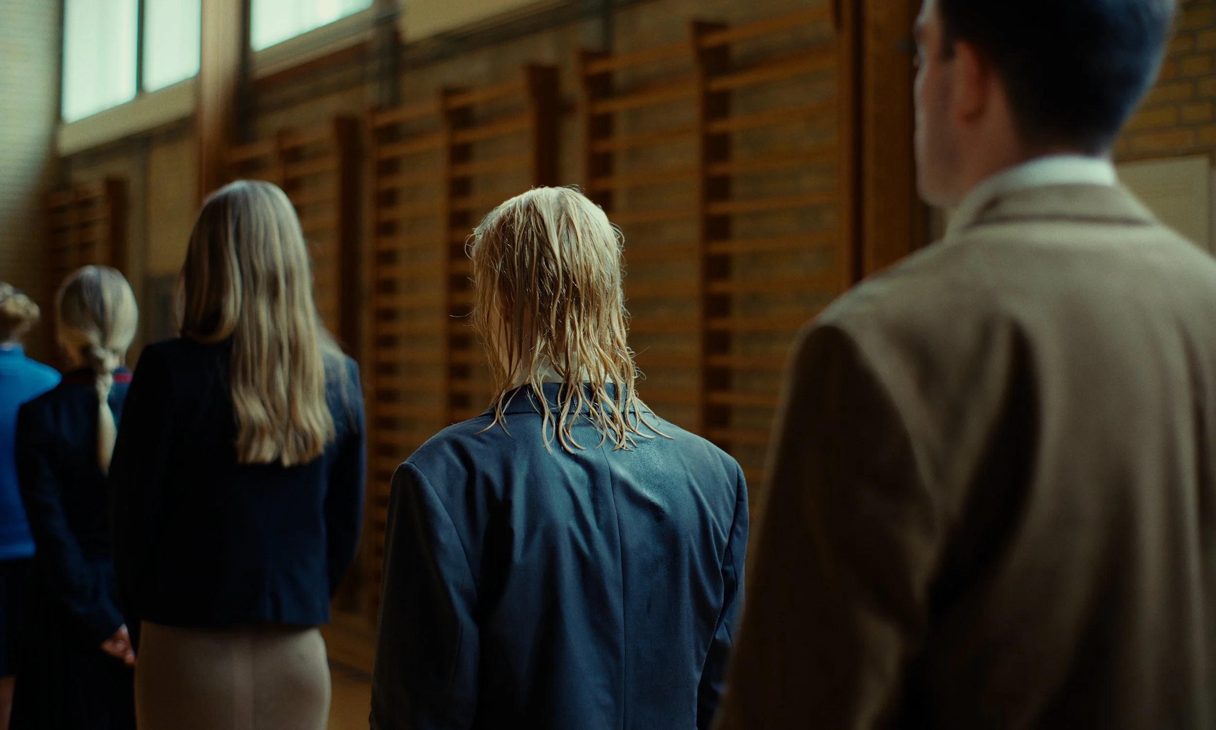 People in formal attire standing in a line indoors, facing away, with a wooden wall in the background.