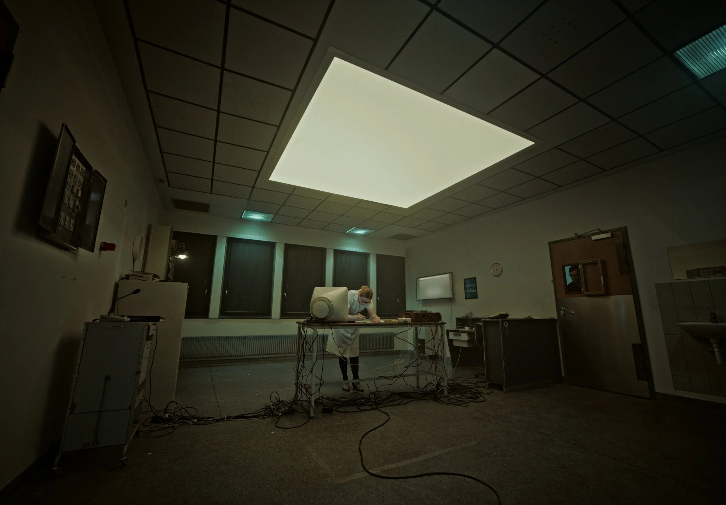A person in a hospital gown and mask working at a messy table with tangled wires in a dimly lit room with a bright ceiling light.
