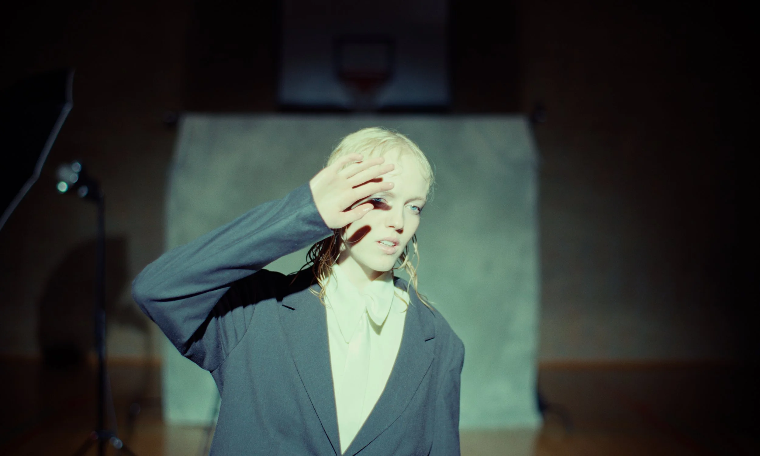 Young woman with blonde hair in a gray blazer with a white shirt, shielding her eyes from the light, in a dimly lit room with photography equipment.