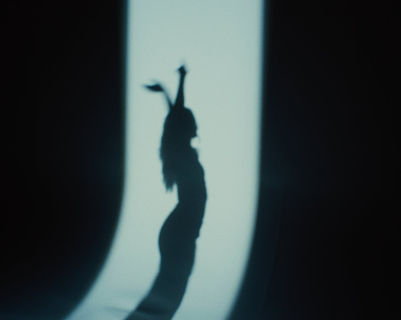 Silhouette of a woman with long hair and bunny ears, with her hands on her hips, standing on her head, creating a bunny shape with her body from a shadow projected on a wall.