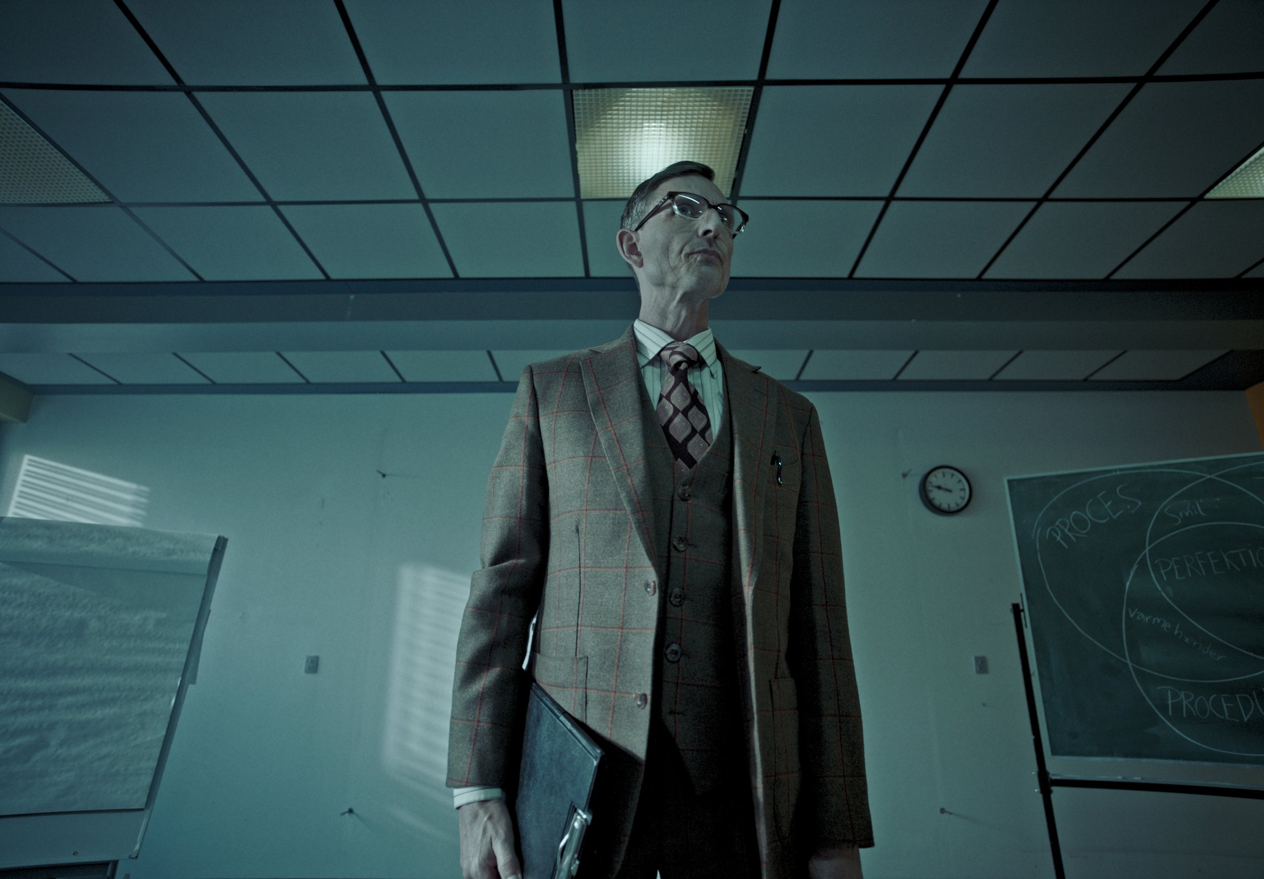 An older man in a brown plaid suit, glasses, and a tie standing in a classroom with a chalkboard and clock in the background.
