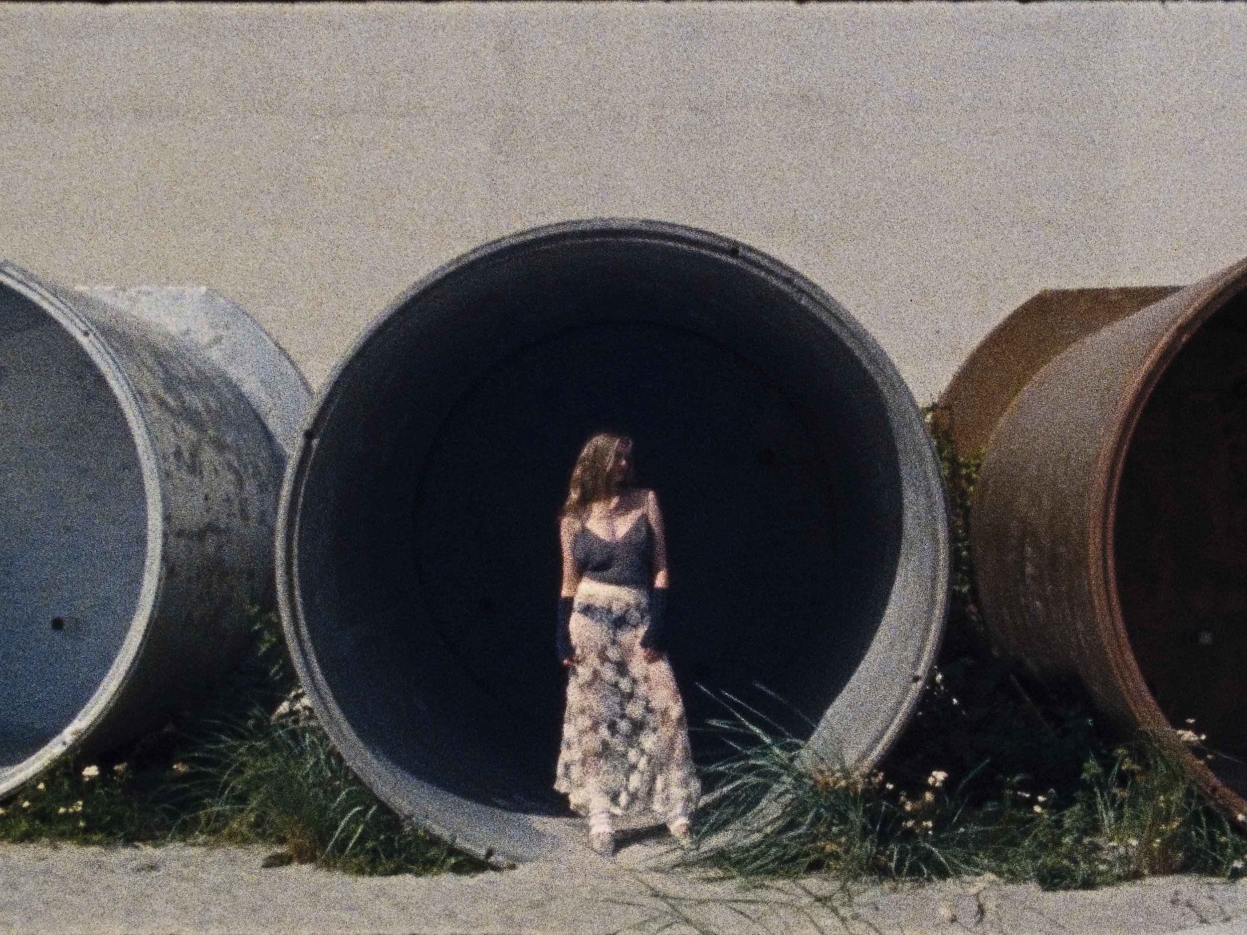 A woman with long wavy hair stands inside a large black metal pipe, wearing a sheer black top and patterned wide-leg pants, with two other large pipes on either side and grass nearby.