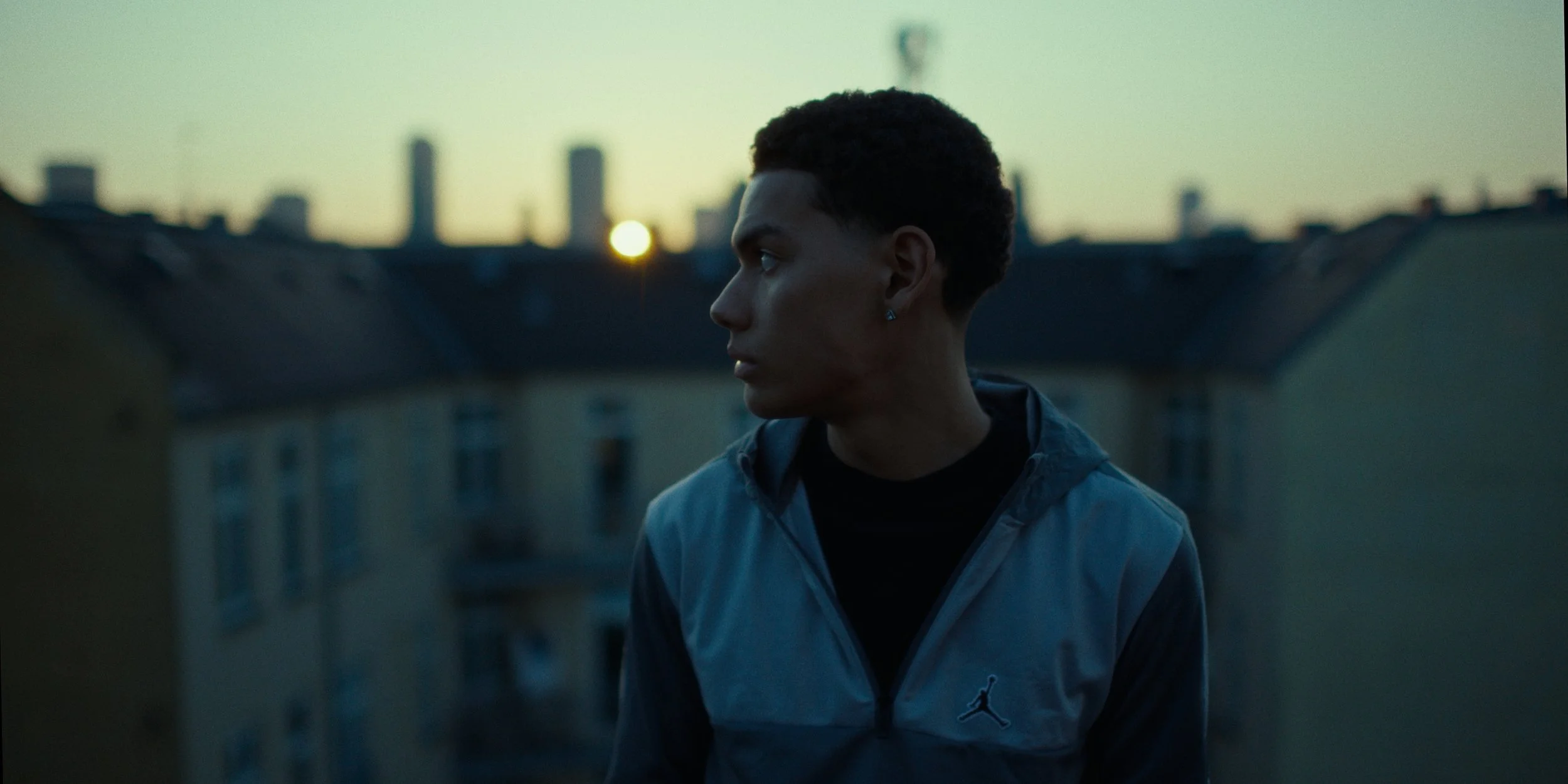A young man with short curly hair wearing a blue and black jacket, standing outdoors on a rooftop at sunset, looking to the side with a city skyline in the background.