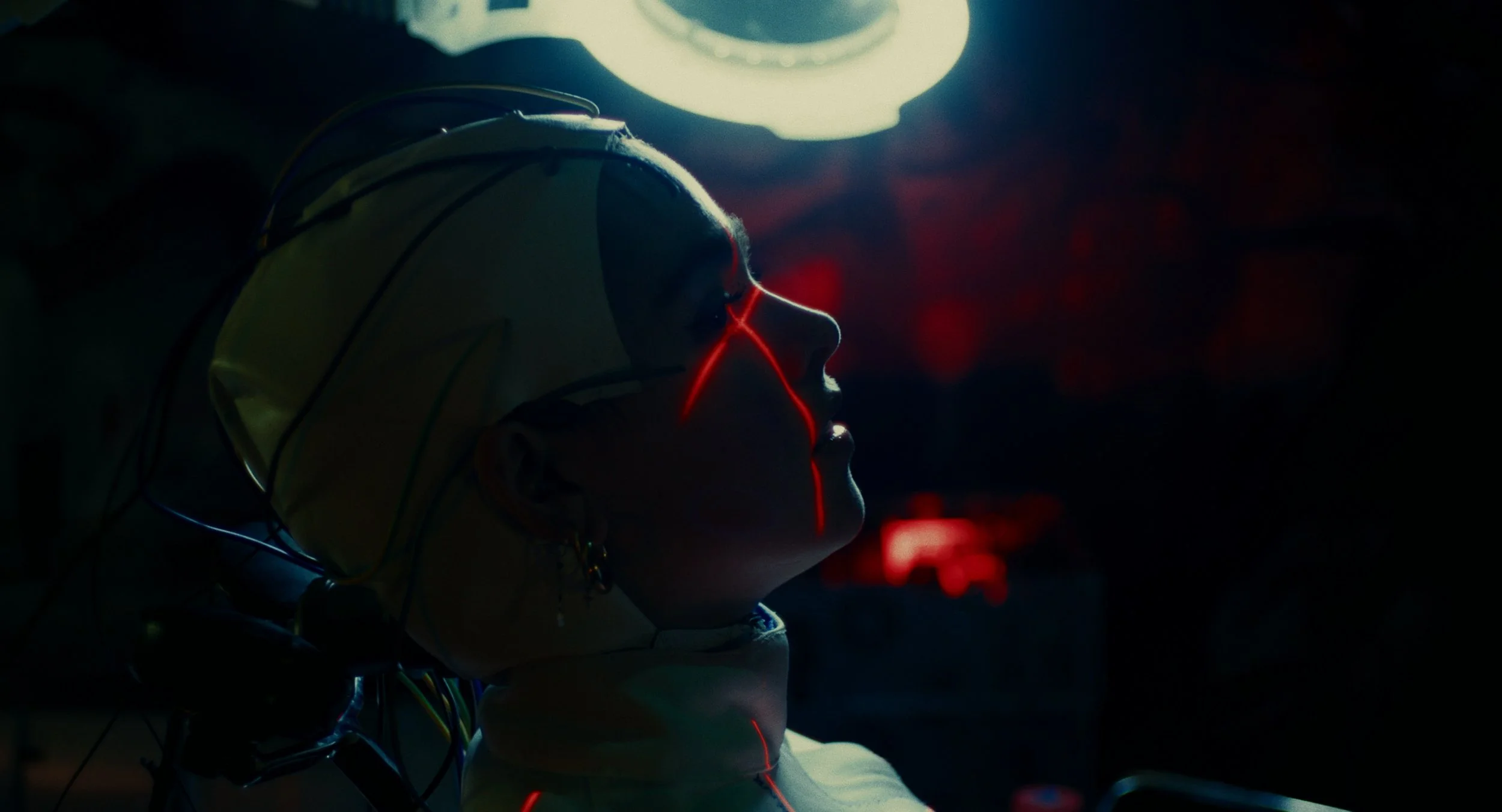 Side profile of a woman with sci-fi or futuristic theme, wearing a helmet with wires and glowing red lines on her face, illuminated by a soft light from above in a dark setting.