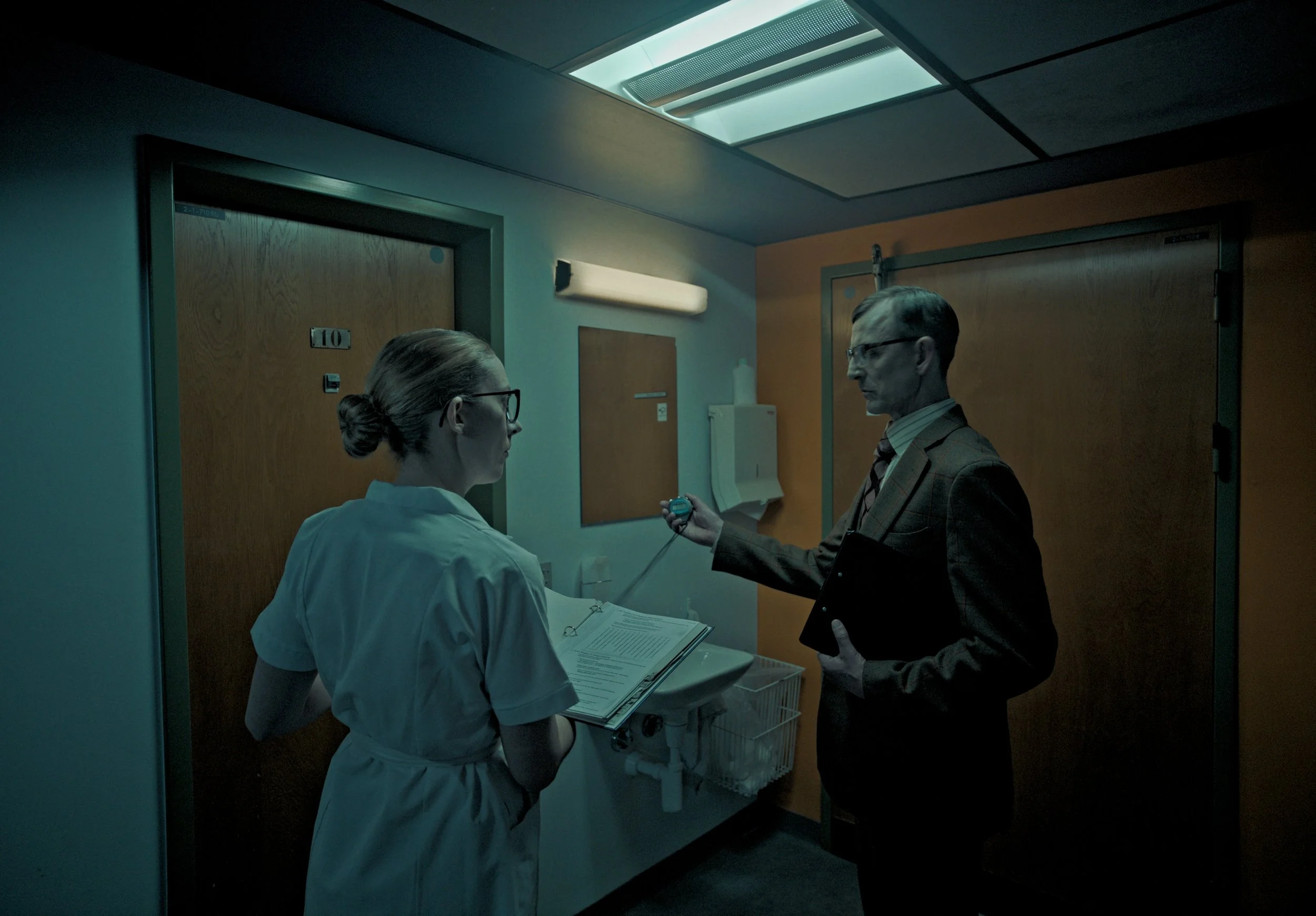 A female nurse with glasses and a bun hairstyle and a male doctor with glasses and a suit in a hospital room. The male doctor is holding a stopwatch or timer, and the nurse is holding a clipboard and clinical documents. The room has wooden doors with