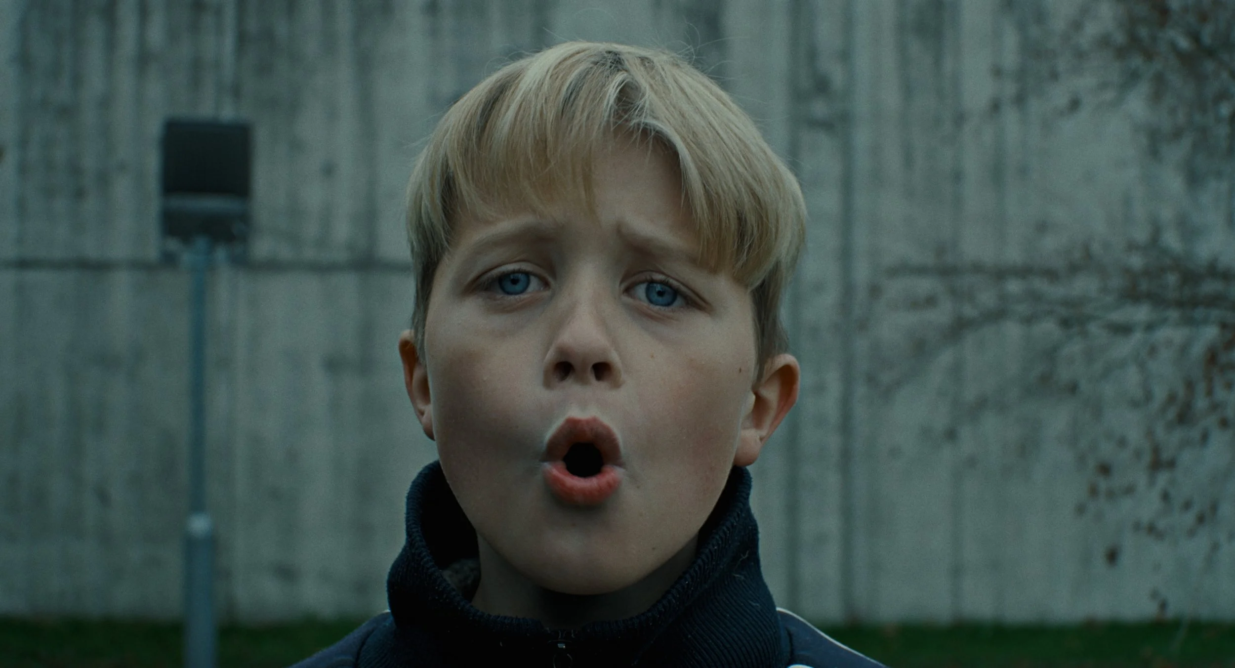 Close-up of a young boy with blond hair and blue eyes, with an expression of surprise or shock, standing outside near a concrete wall.