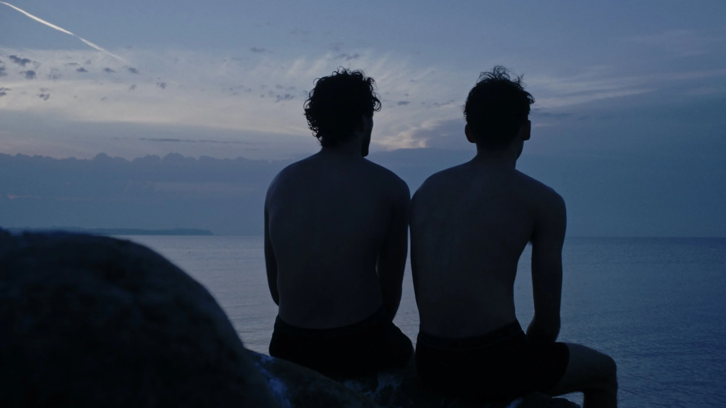 Two shirtless young men sitting on a large rock by the water at dusk, looking out at the ocean with cloudy sky above.