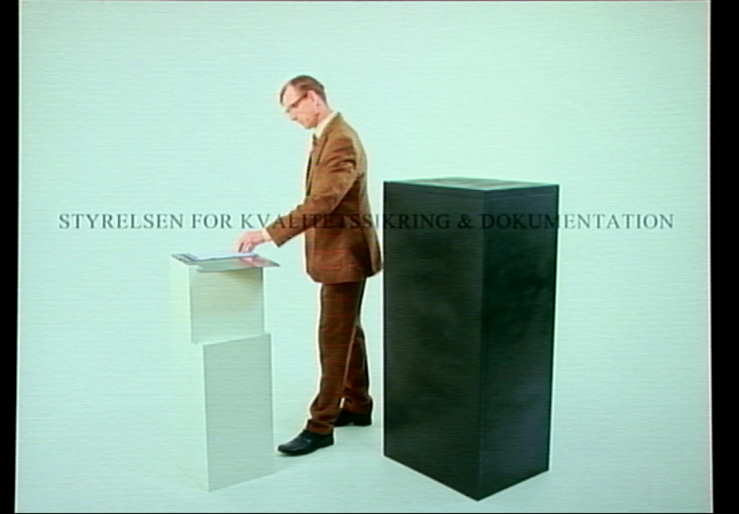 A man in a brown suit and glasses standing by two pedestals, with the larger black one on the right and a smaller light-colored one on the left, with Norwegian text overlayed.